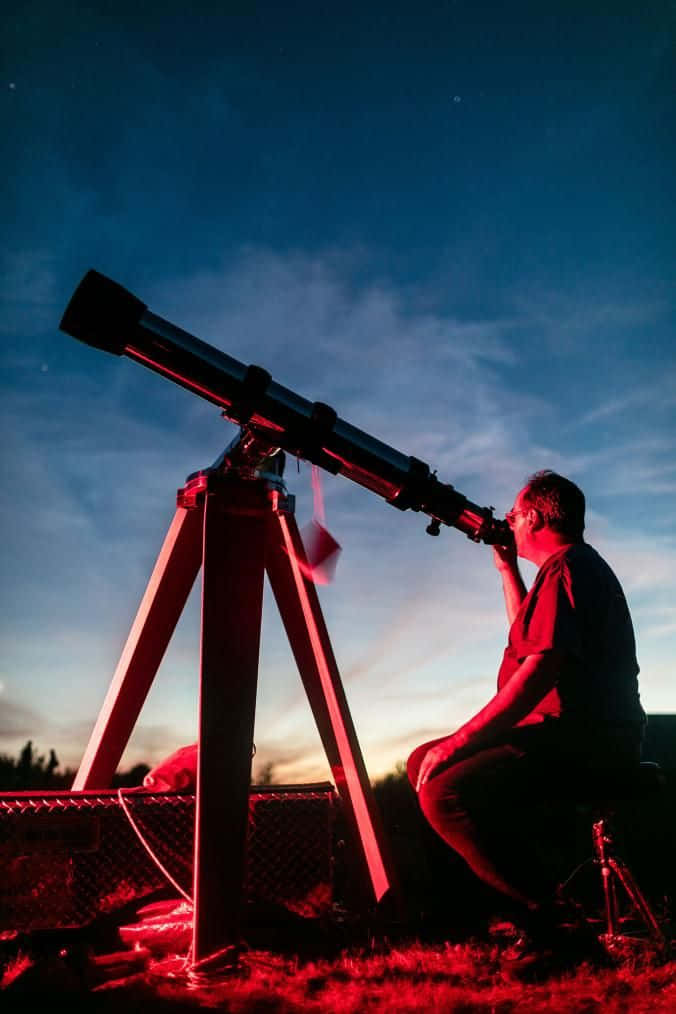Telescope 桌布 壁紙 — 一個剪影人物跪在一個沐浴在紅光中的望遠鏡旁，背景是暮色天空，星星開始浮現。