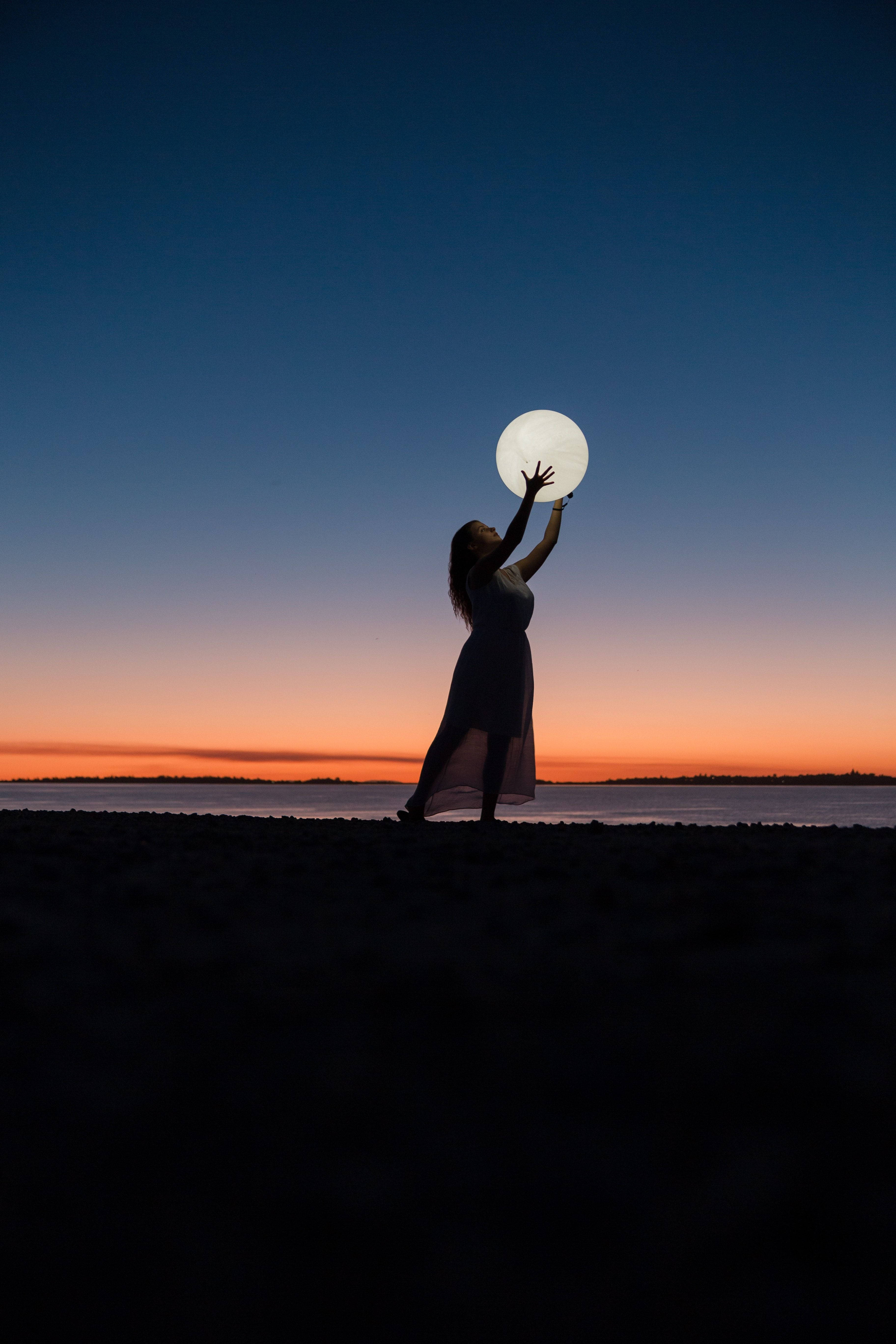月夜星空 桌布 壁紙 — 一個剪影女子在黃昏時站在沙灘上，伸手觸碰一個發光的球體，背景是橙色和深藍色的漸變天空。