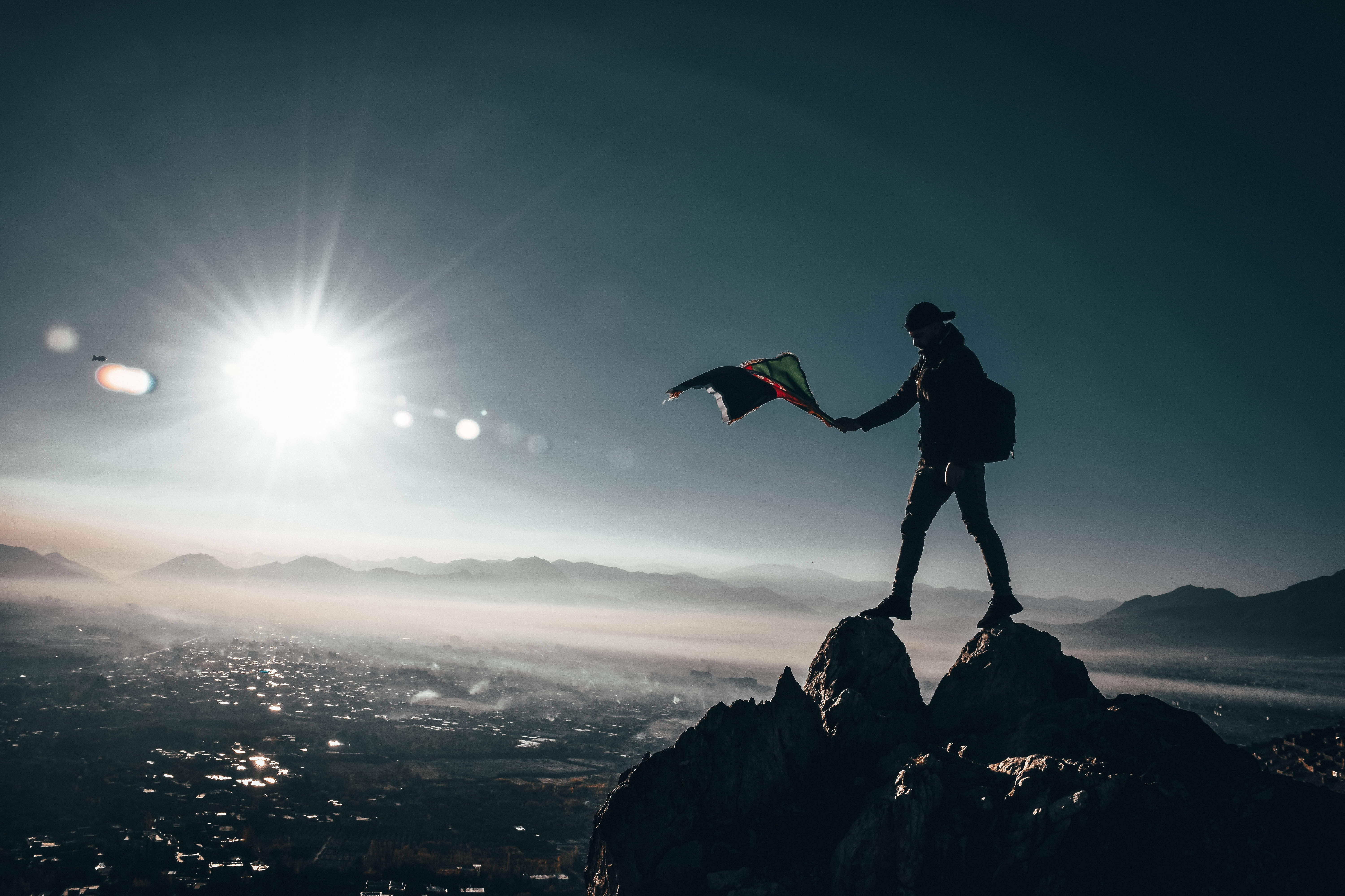 Brave 桌布 壁紙 — 一個剪影登山者站在岩石山頂上，在日出時舉著旗幟，俯瞰下方霧氣瀰漫的山谷。