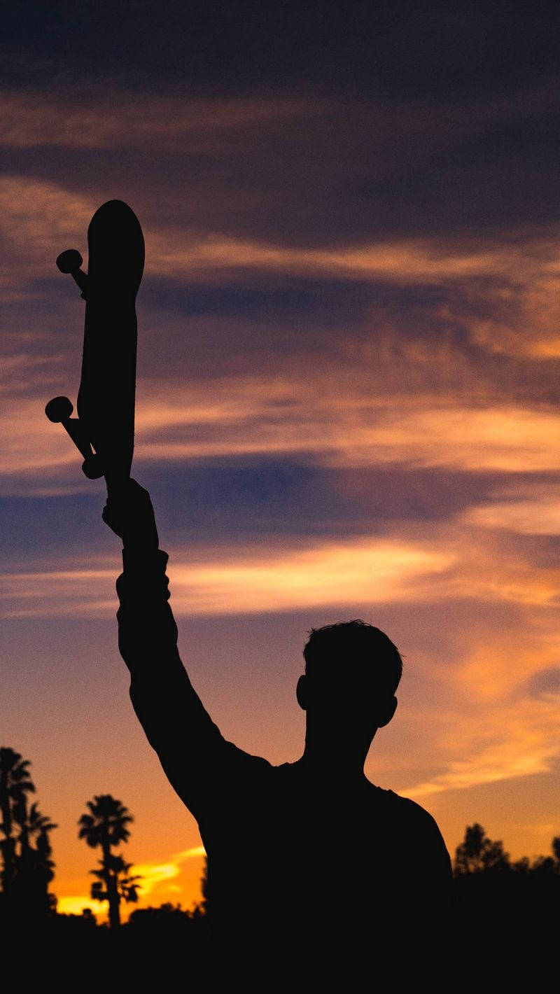 Silhouette 桌布 壁紙 — 一個剪影的滑板手勝利地高舉滑板，背景是充滿活力的日落天空，下方有棕櫚樹。