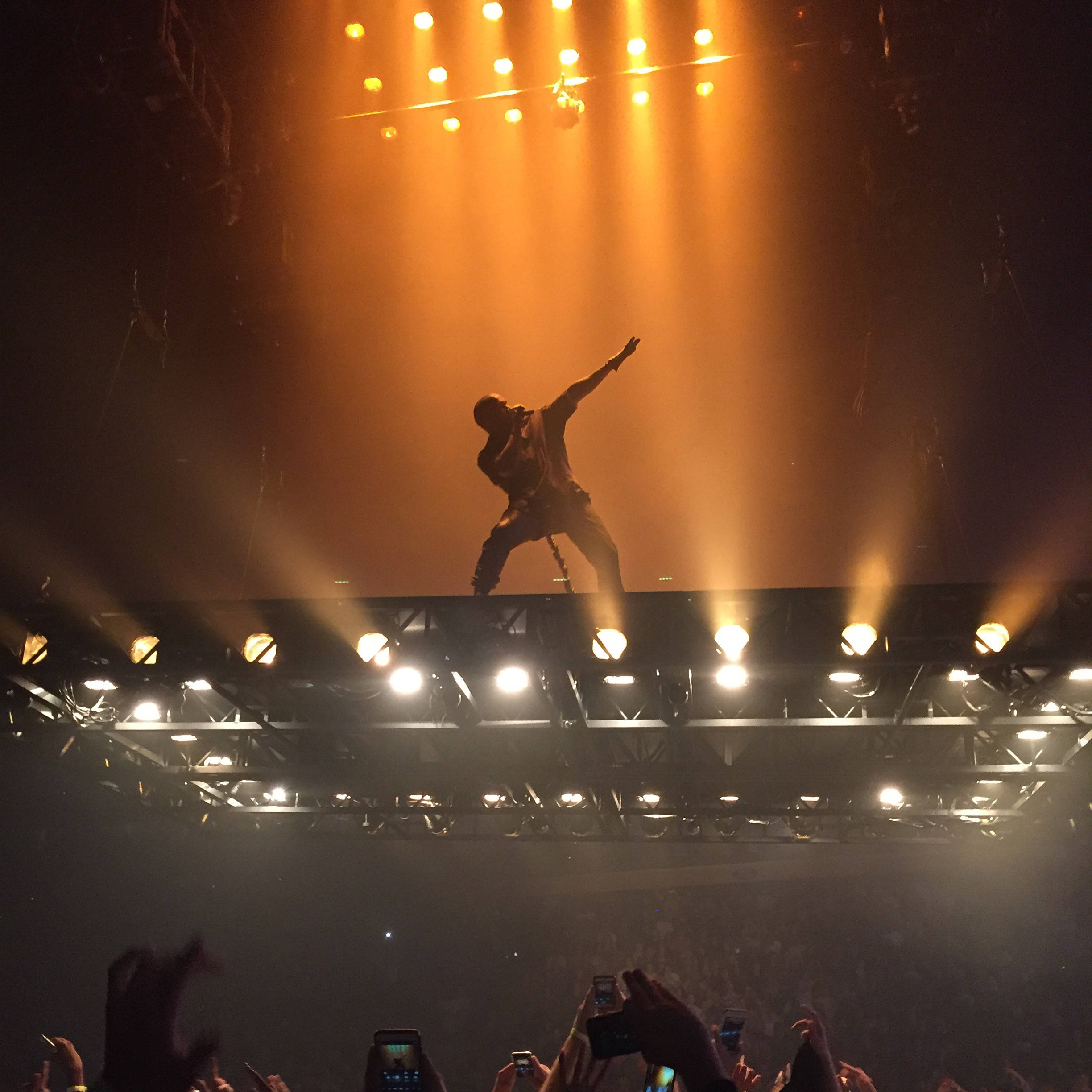 Kanye 桌布 壁紙 — 一個剪影表演者在燈光明亮的舞台上跳躍，而下面的粉絲舉起手機捕捉這個電光火石的演唱會時刻。