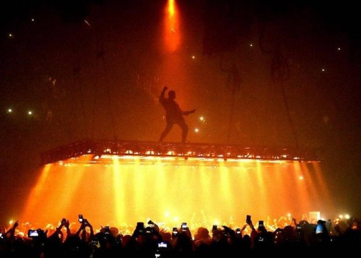 Kanye 桌布 壁紙 — 一個剪影表演者在發光的舞台上跳舞，而下面的群眾舉起手機捕捉充滿活力的橙色演唱會燈光。