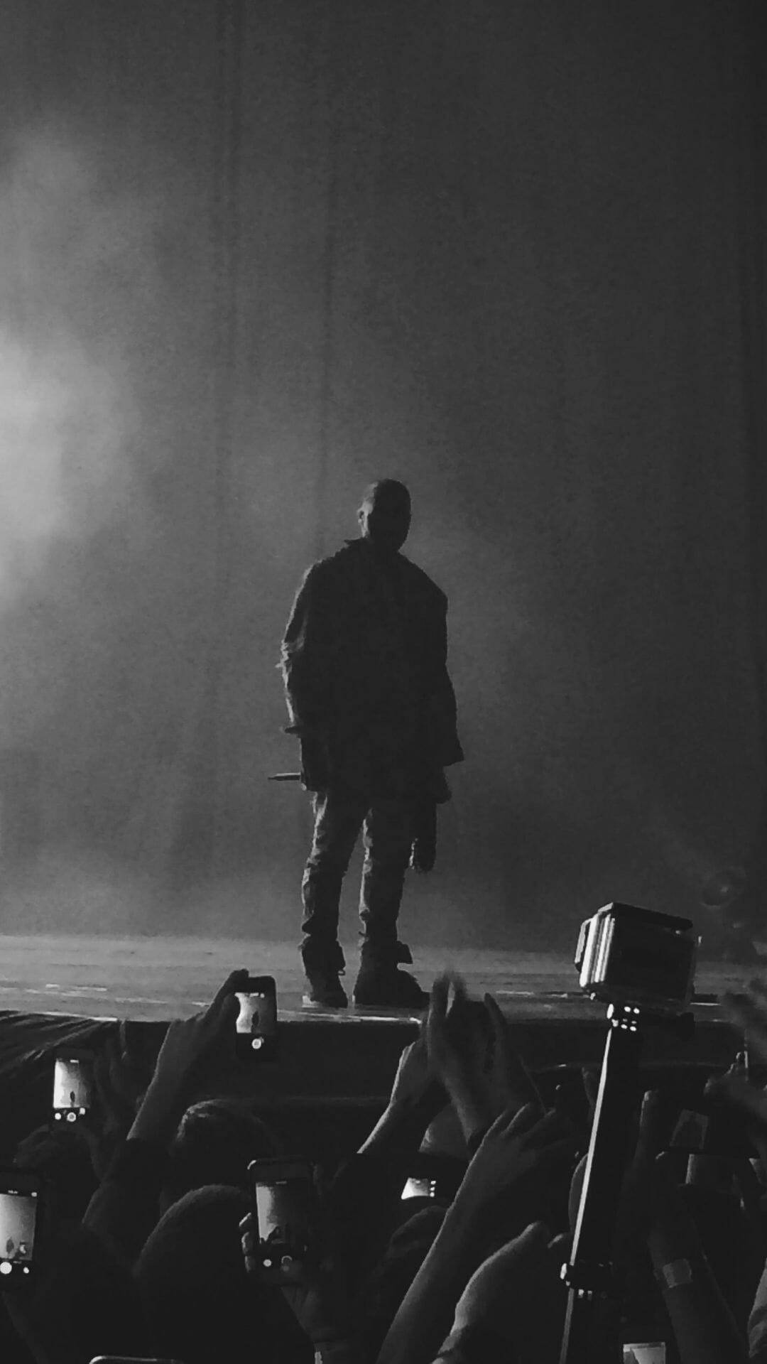 Kanye 桌布 壁紙 — 一個剪影表演者獨自站在煙霧瀰漫的舞台上，人群的手舉起手機捕捉這一刻。