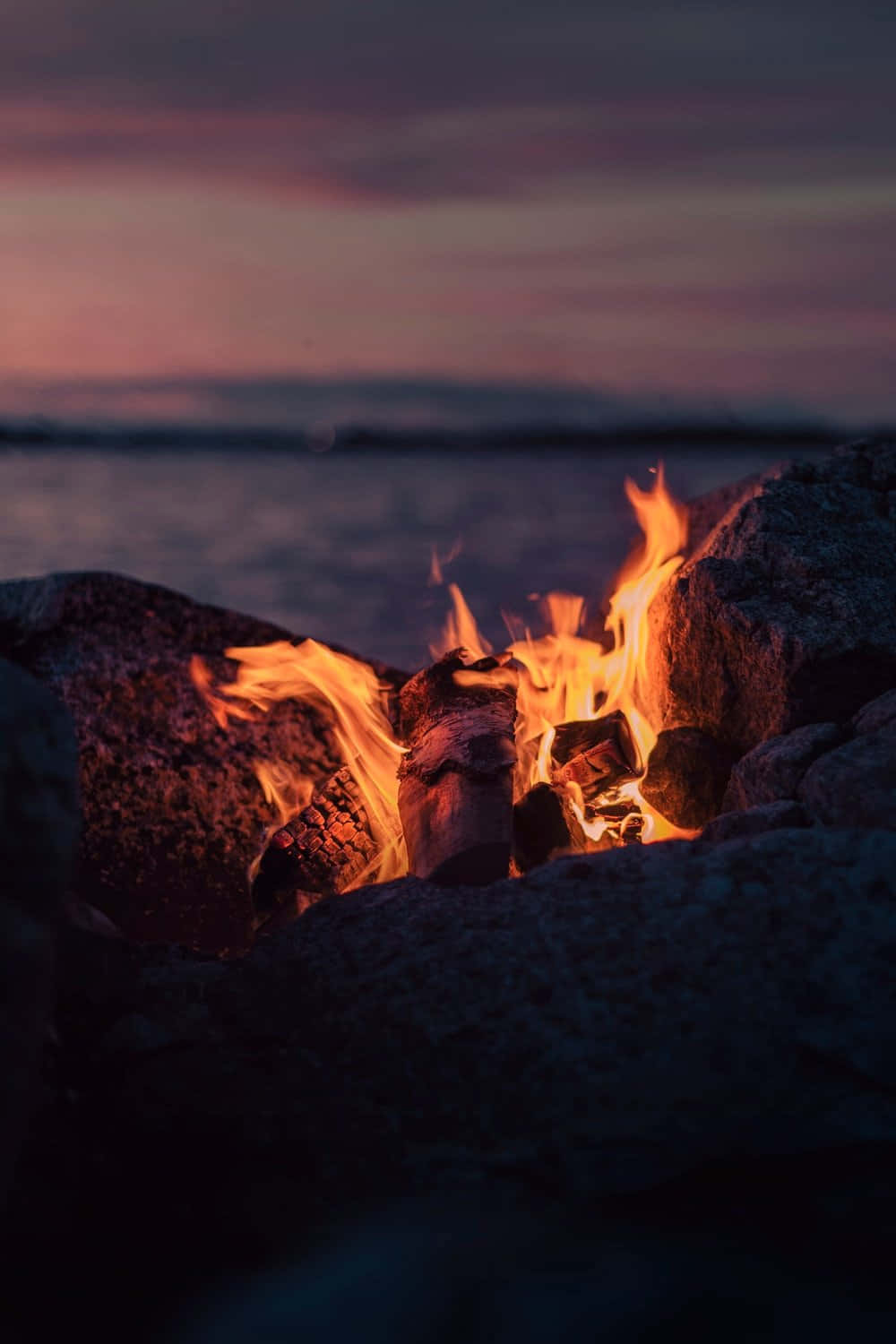 Cozy Fire 桌布 壁紙 — 一個劈啪作響的營火在黑暗的岩石間明亮地燃燒，旁邊是一個平靜的湖泊，上方是塗上粉紅色和紫色色調的暮色天空。