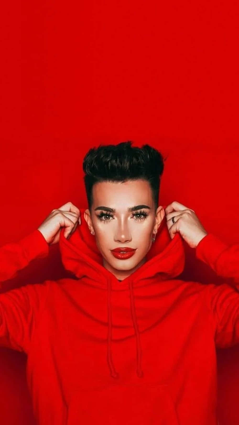 James Charles 桌布 壁紙 — 一個化著大膽妝容、留著黑色短髮的人，在純紅色背景前拉動一件鮮紅色衛衣的帽子。