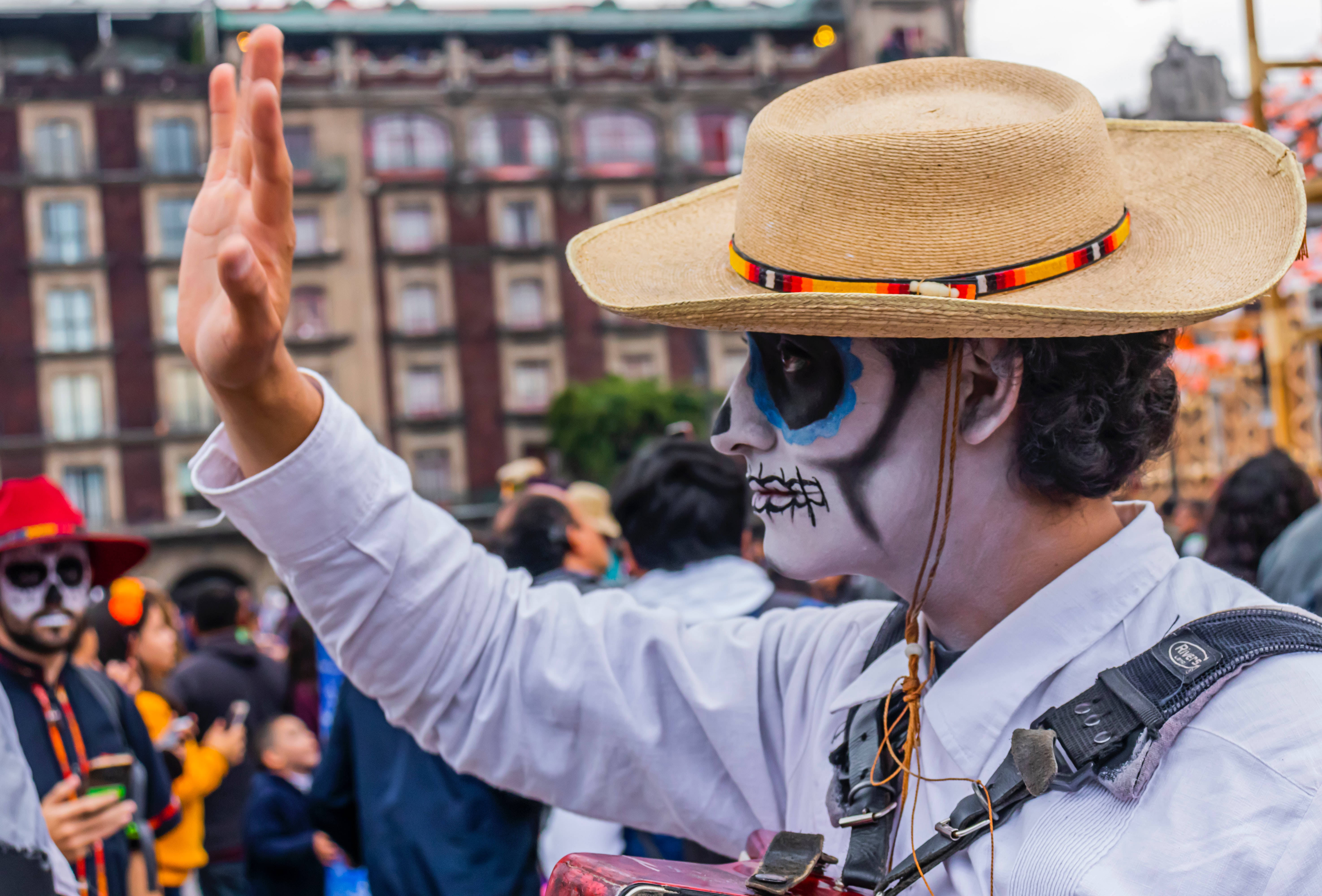 Mexican iPhone 桌布 壁紙 — 一個化著糖骷髏妝、戴著草帽的人，在歷史建築附近擁擠的街頭節日上揮手。