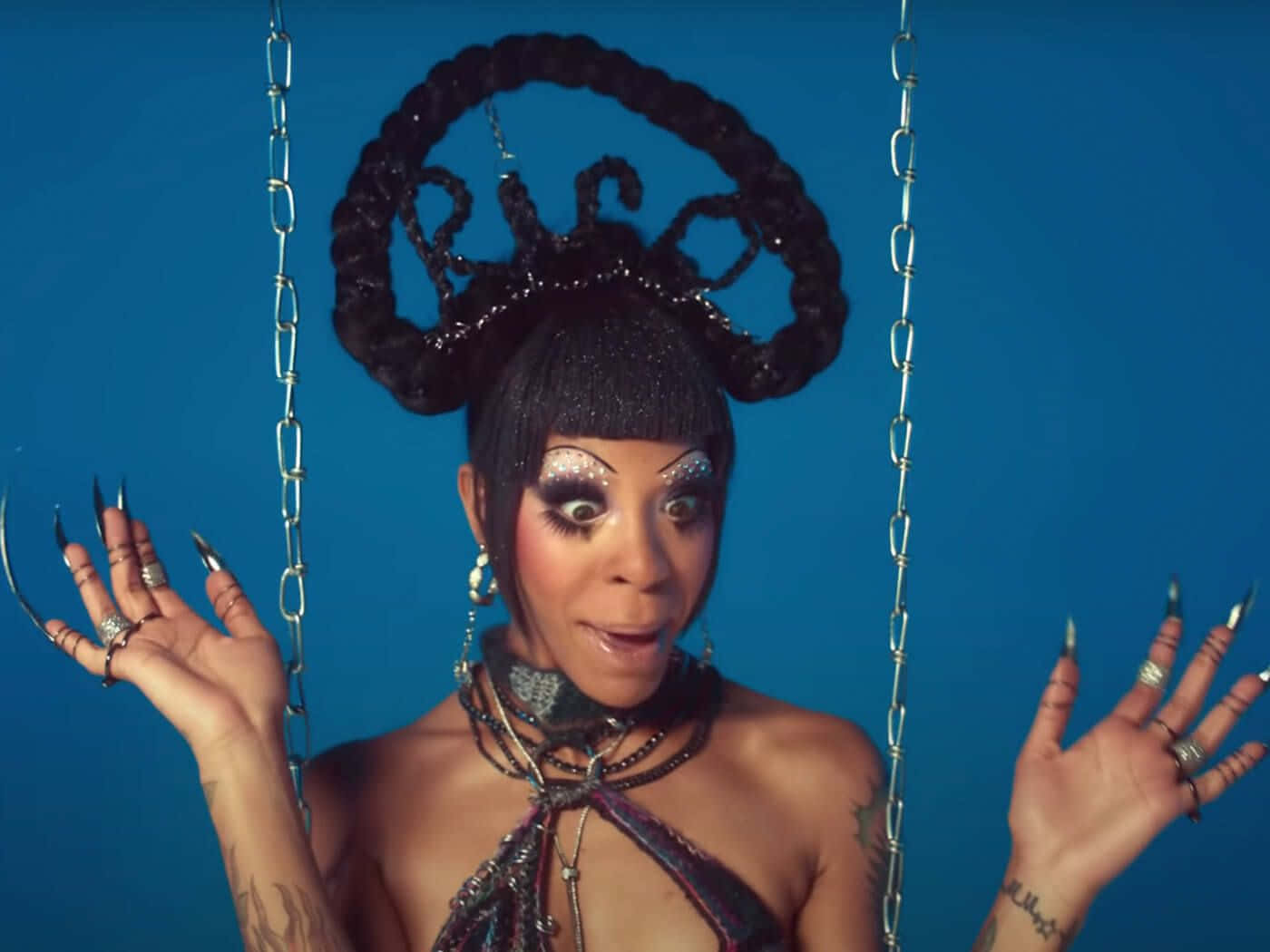 Rico Nasty 桌布 壁紙 — 一個化著閃亮妝容、戴著華麗辮子頭飾的人，被鎖鏈懸掛在鮮豔的藍色背景前。