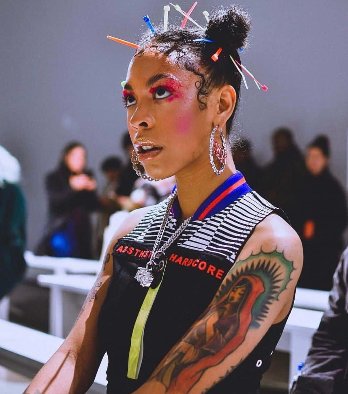 Rico Nasty 桌布 壁紙 — 一個化著鮮豔妝容、戴著彩色髮夾的人，展示著有刺青的手臂和大膽的街頭服飾，目光強烈。