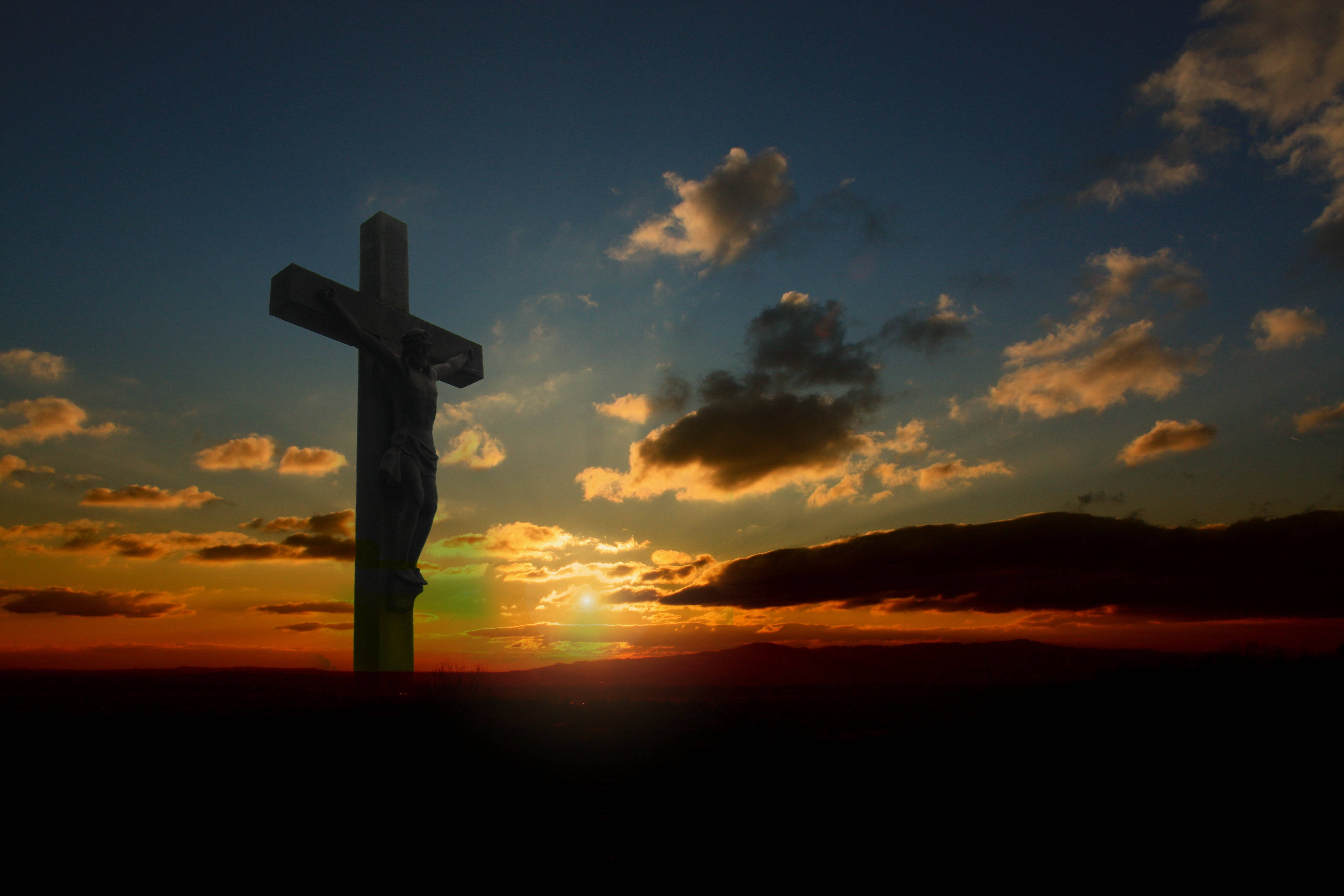 Christ 桌布 壁紙 — 一個十字架雕像在戲劇性的日落天空下呈現剪影，天空有金色雲彩和深藍色調。