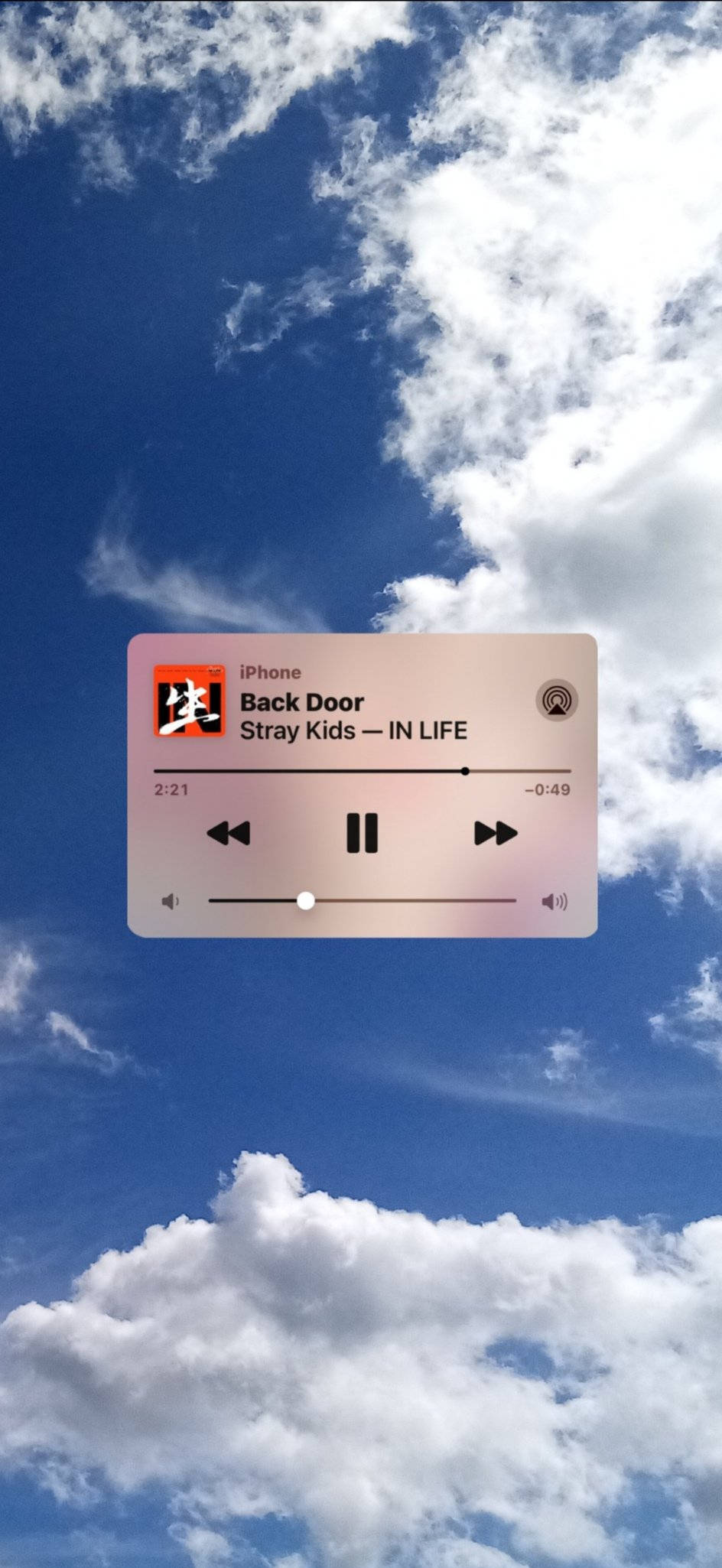 音樂 桌布 壁紙 — 一個半透明的iPhone音樂播放器疊加層顯示Stray Kids的歌曲Back Door，背景是充滿活力的藍天和蓬鬆的白雲。