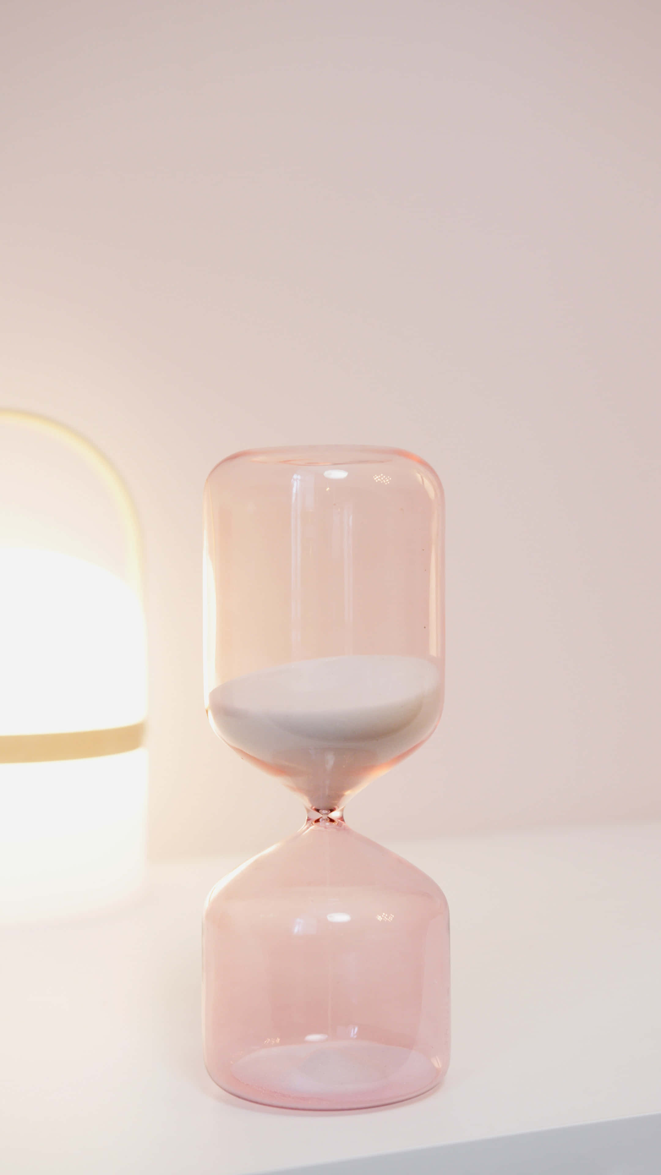 Pastel Vintage iPhone 桌布 壁紙 — 一個半透明粉紅色沙漏，內有白色沙粒，放在白色表面上，旁邊有一盞發光的燈，背景是柔和的粉紅色牆壁。