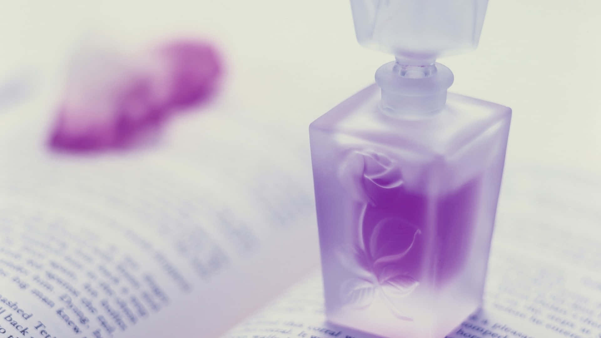 Floral Fragrance 桌布 壁紙 — 一個半透明紫色香水瓶，帶有花卉蝕刻圖案，放在一本打開的書上，背景有一個柔和焦點的紫色心形物體。