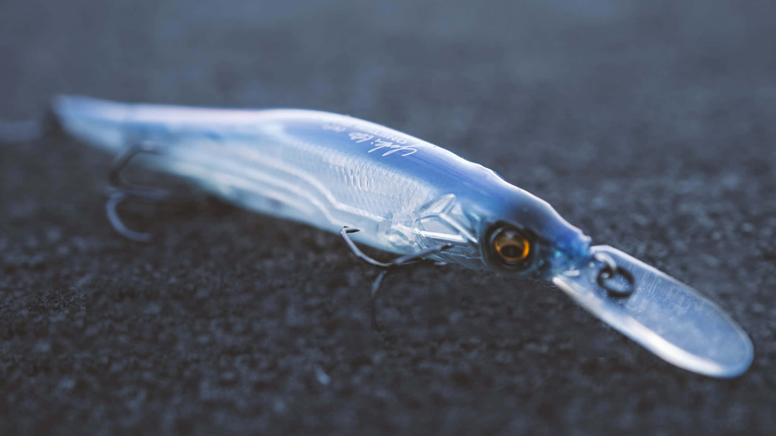 Fishing Bait 桌布 壁紙 — 一個半透明藍色魚餌，帶有橙色眼睛和銀色魚鉤，放置在深色紋理地面上。