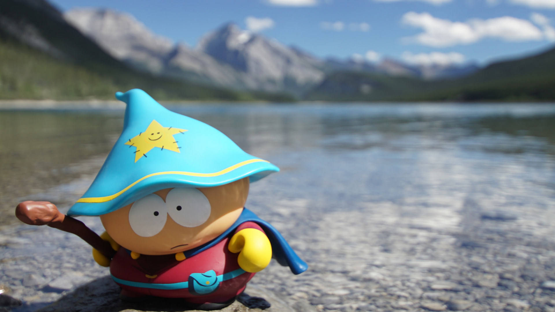 Cartman 桌布 壁紙 — 一個南方公園巫師人偶站在岩石湖岸上，背景是白雪覆蓋的山脈和藍天。