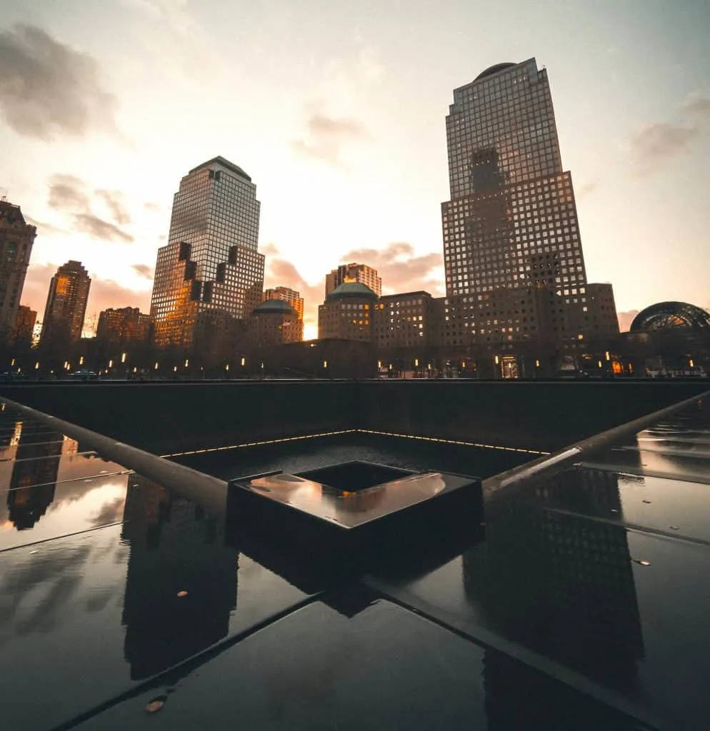 911 Memorial 桌布 壁紙 — 一個反射池映照出高聳的摩天大樓，在溫暖的日落天空下，伴隨著柔軟的雲朵。
