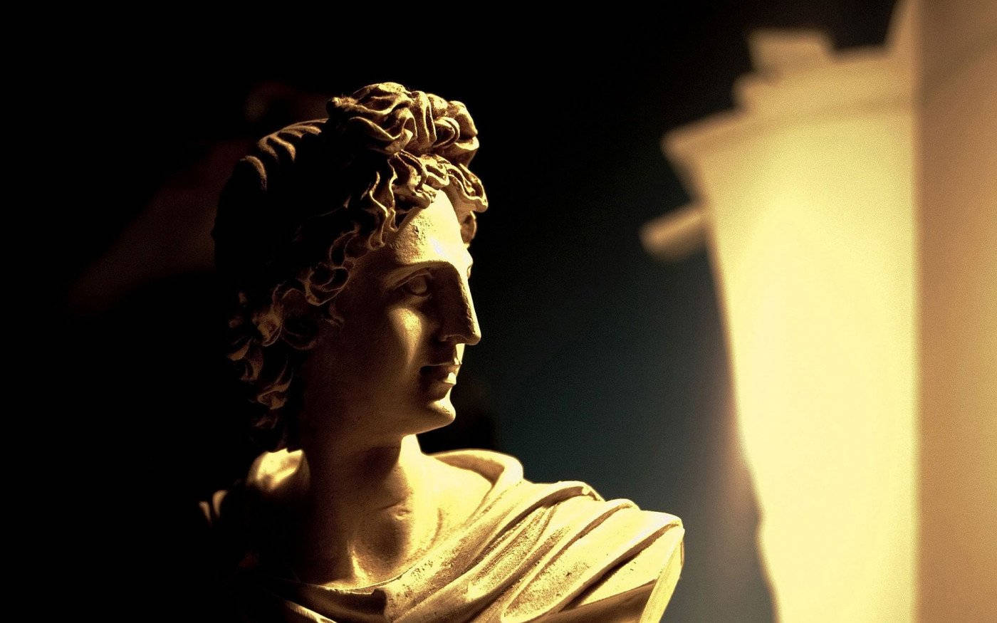 Statue Aesthetic 桌布 壁紙 — 一個古典大理石半身像，描繪一個捲髮的年輕人物，在黑暗背景中戲劇性地被照亮，旁邊有一根溫暖發光的柱子。