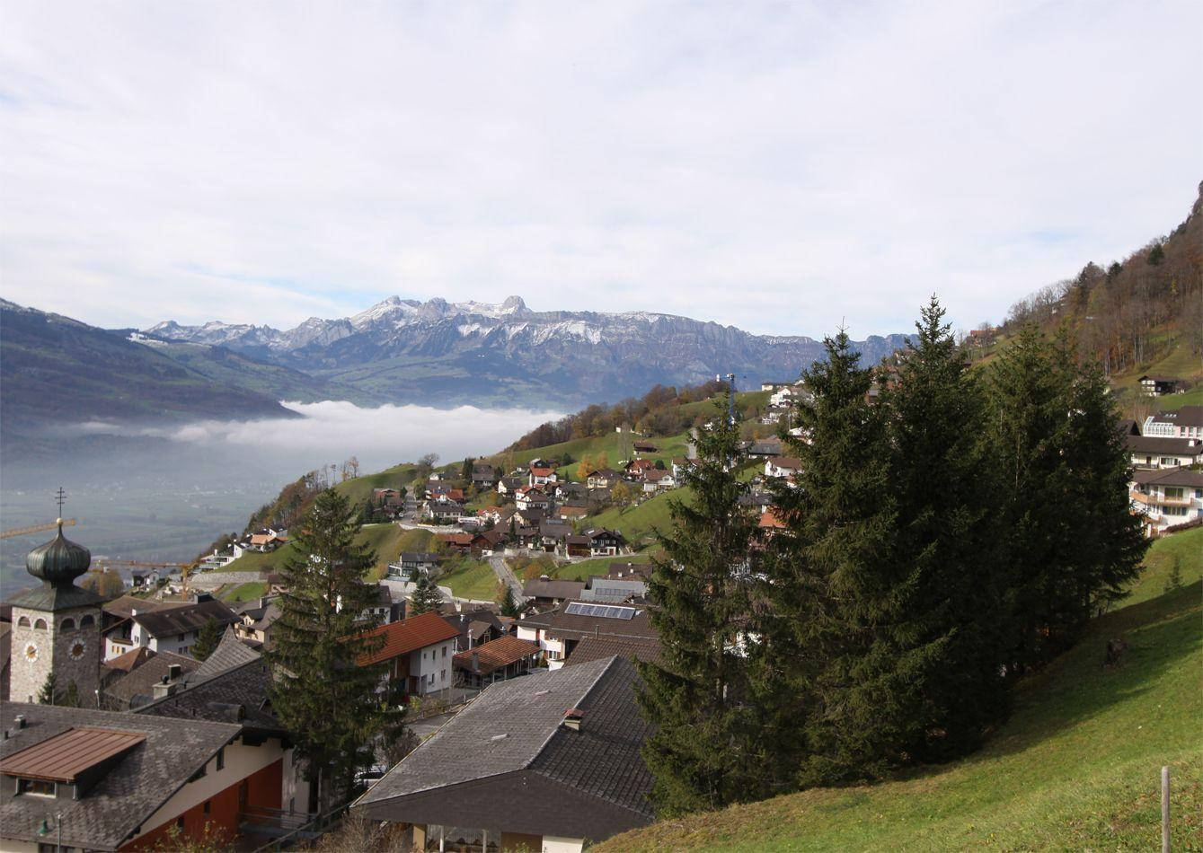 Liechtenstein 桌布 壁紙 — 一個古雅的阿爾卑斯山村依附在長滿青草的山坡上，俯瞰著霧氣瀰漫的湖泊，遠處是白雪覆蓋的山峰。