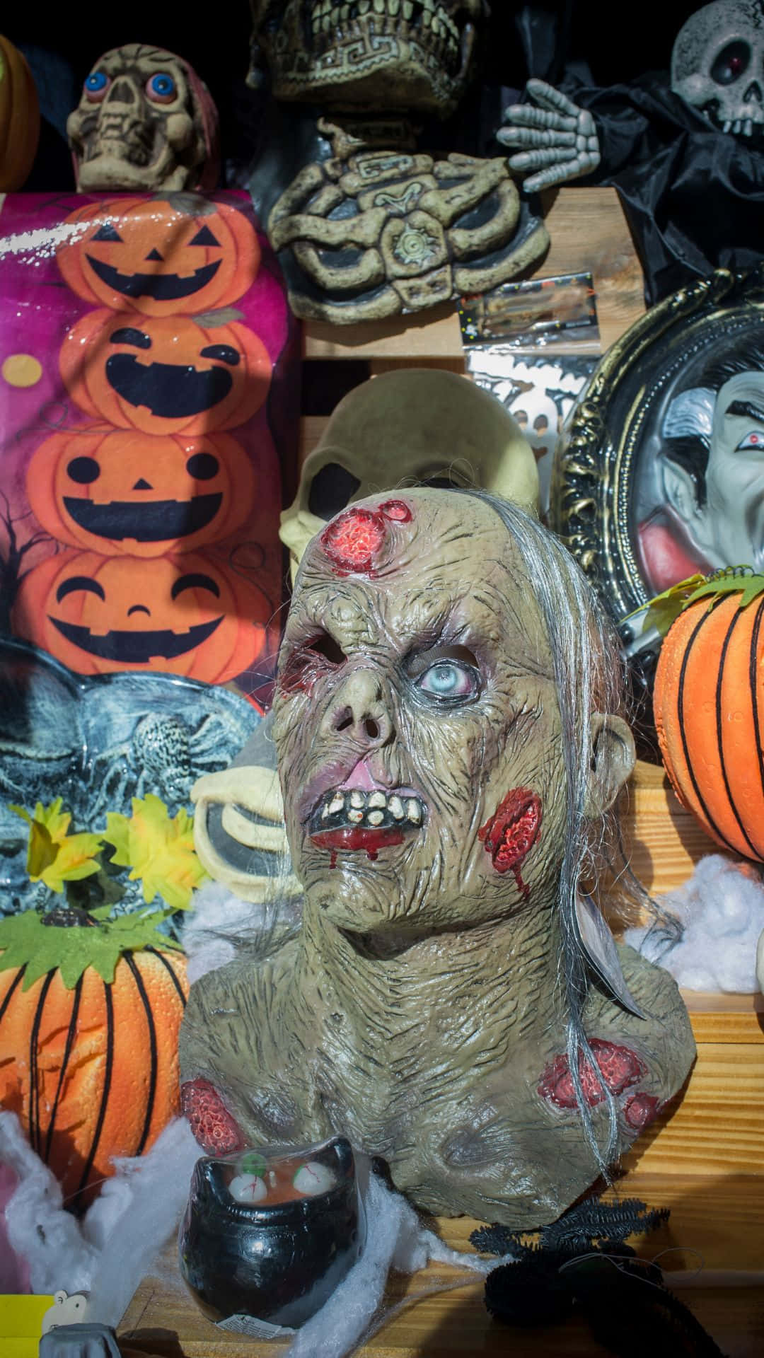 Horror Masks 桌布 壁紙 — 一個可怕的殭屍半身像，帶有血淋淋的傷口和發光的眼睛，坐在萬聖節裝飾品（如南瓜燈和頭骨）的中心。