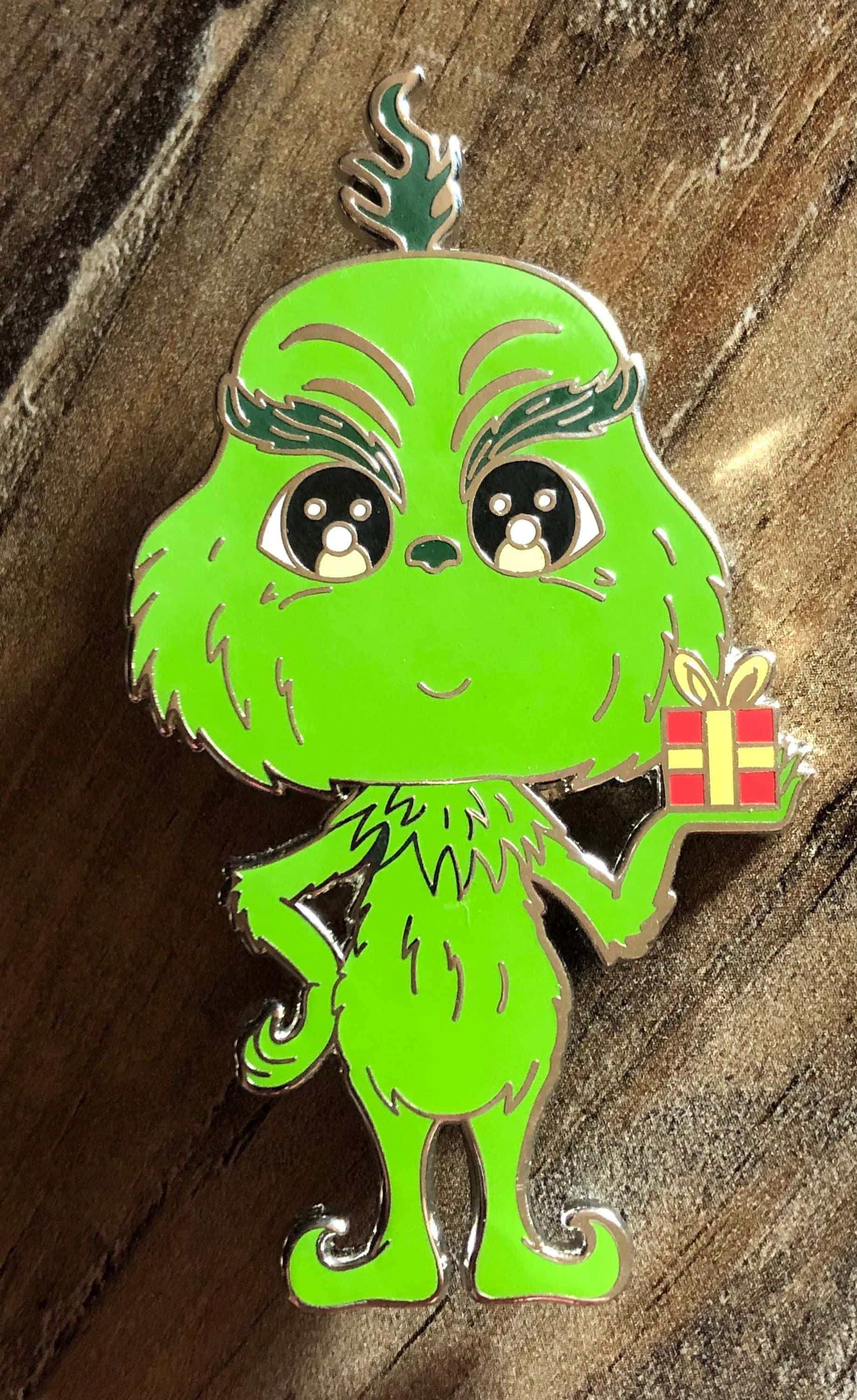 Cute Grinch 桌布 壁紙 — 一個可愛的Q版綠色鬼靈精琺瑯別針拿著禮物，放在質樸的木質表面上。