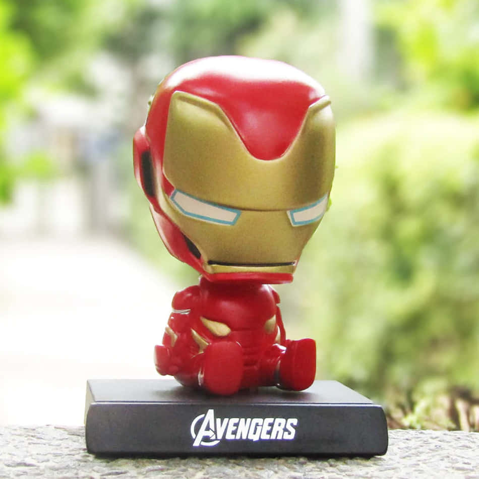 Iron Man Bobbleheads 桌布 壁紙 — 一個可愛的Q版鋼鐵人公仔坐在戶外的復仇者聯盟基地上，背後有模糊的綠色樹葉。