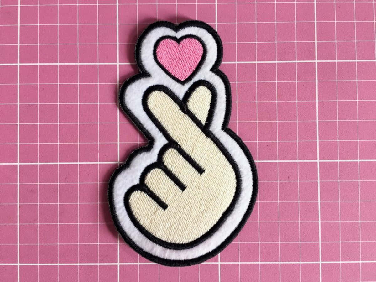 Finger Heart 桌布 壁紙 — 一個可愛的刺繡補丁，一隻手做出心形手勢，上方有一個粉紅色心形，背景是粉紅色網格。
