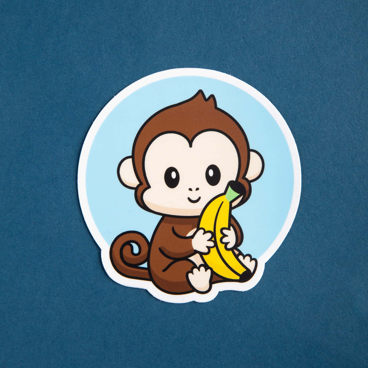 Monkey 桌布 壁紙 — 一個可愛的卡通猴子貼紙，拿著香蕉，背景為藍色。