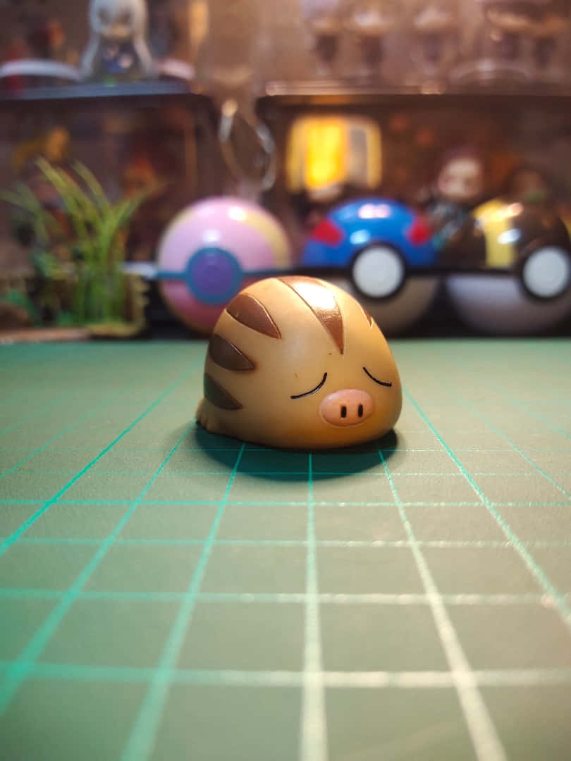 Pokémon 手機 桌布 壁紙 — 一個可愛的棕褐色和棕色豬狀寶可夢公仔靜靜地躺在綠色網格墊上，背景有模糊的寶可夢球玩具。