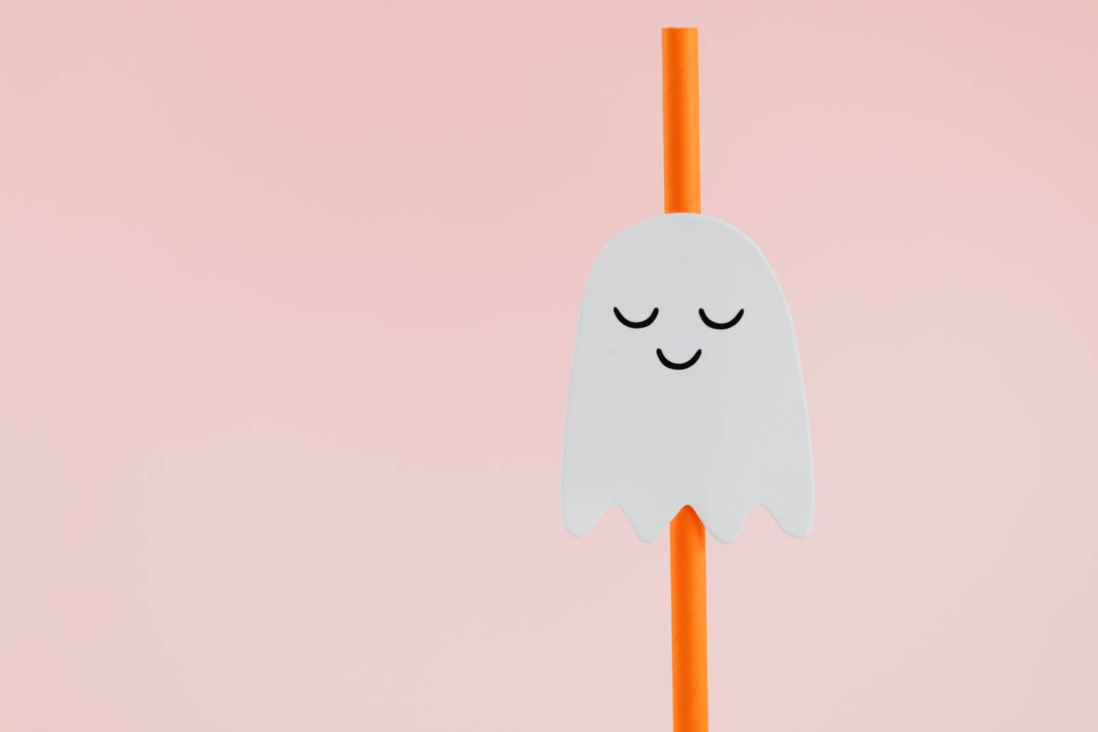 Cute Halloween Desktop 桌布 壁紙 — 一個可愛的白色幽靈形狀吸管蓋，帶有微笑的臉，放在橙色吸管上，背景是柔和的粉紅色。
