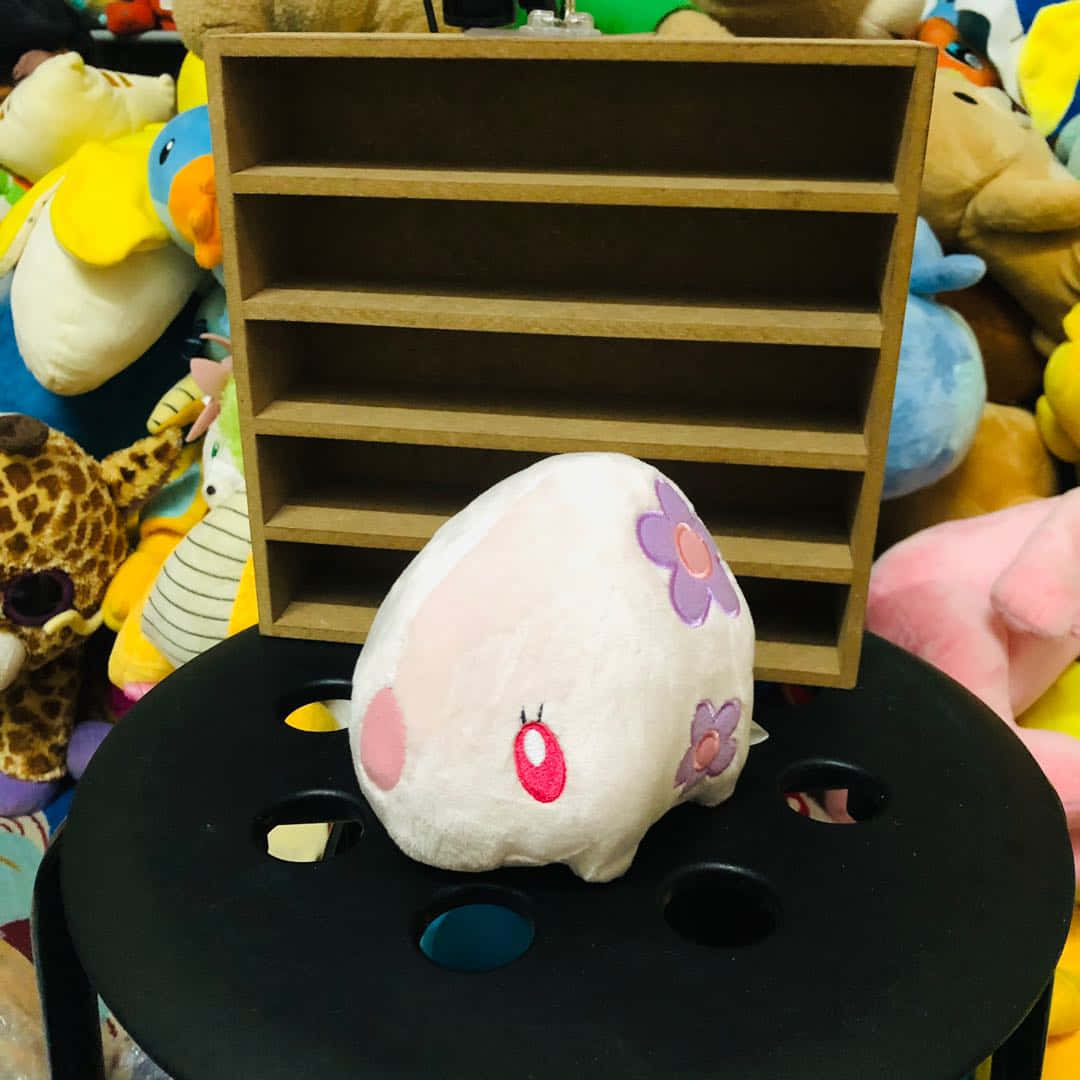 Pokémon 手機 桌布 壁紙 — 一個可愛的白色毛絨玩具，粉紅臉頰和紫色花朵，坐在黑色凳子上，周圍環繞著色彩繽紛的填充動物。