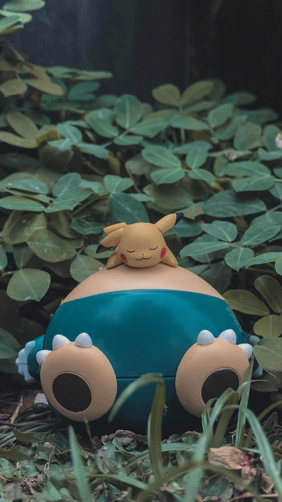 Pokemon Toys 桌布 壁紙 — 一個可愛的皮卡丘公仔安詳地躺在一個青綠色的寶可夢球上面，周圍是茂密的綠色植物。