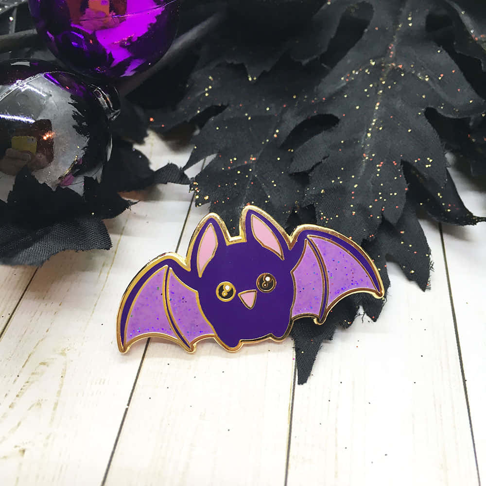 Bats 桌布 壁紙 — 一個可愛的紫色閃光蝙蝠別針，帶有金色邊飾，放在白色木頭上，旁邊有黑色閃光布料和紫色裝飾品。