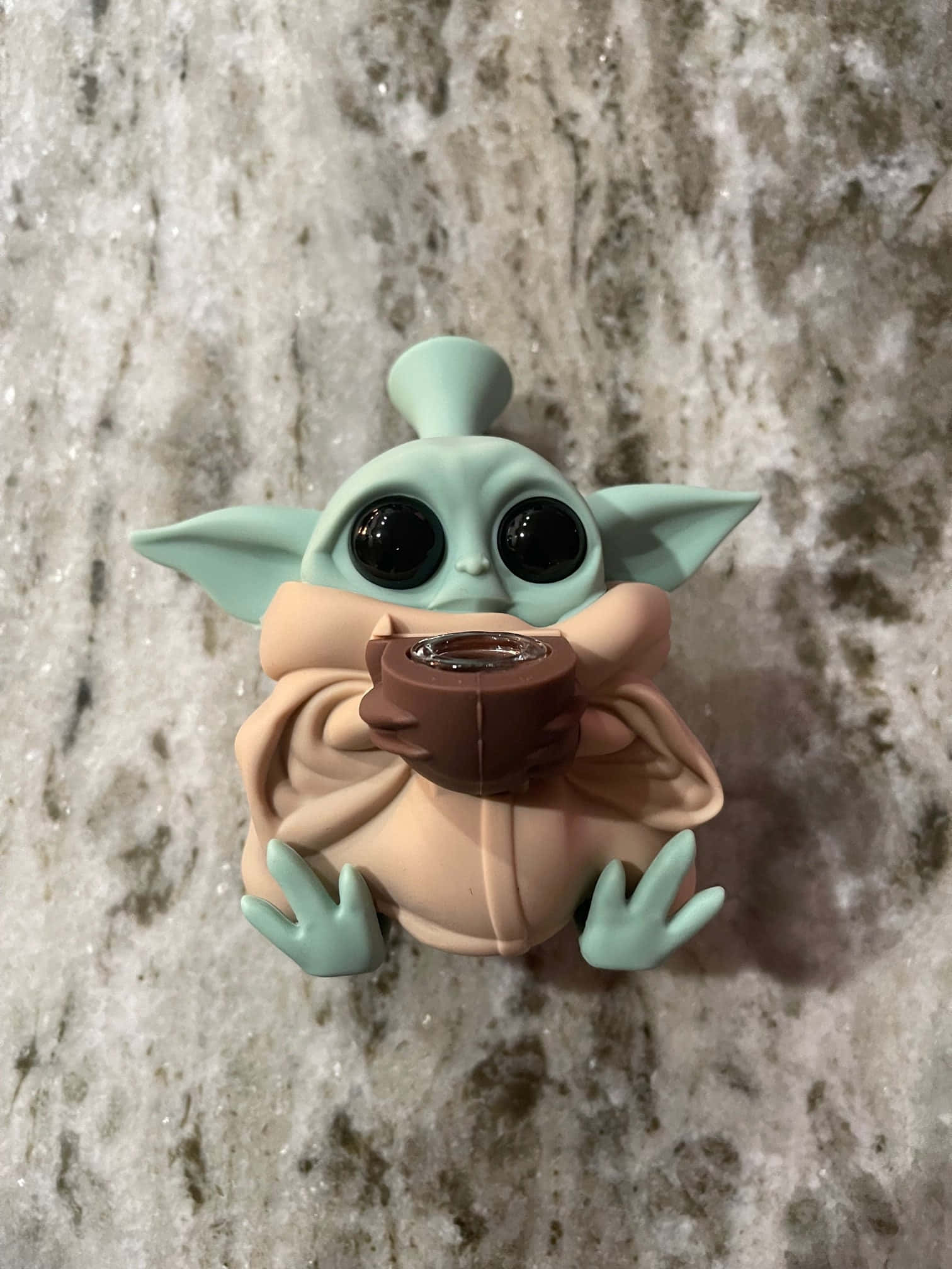 Baby Yoda Aesthetic 桌布 壁紙 — 一個可愛的綠色尤達寶寶公仔，身穿米色長袍，在帶有斑點的灰色檯面上拿著一個棕色杯子。