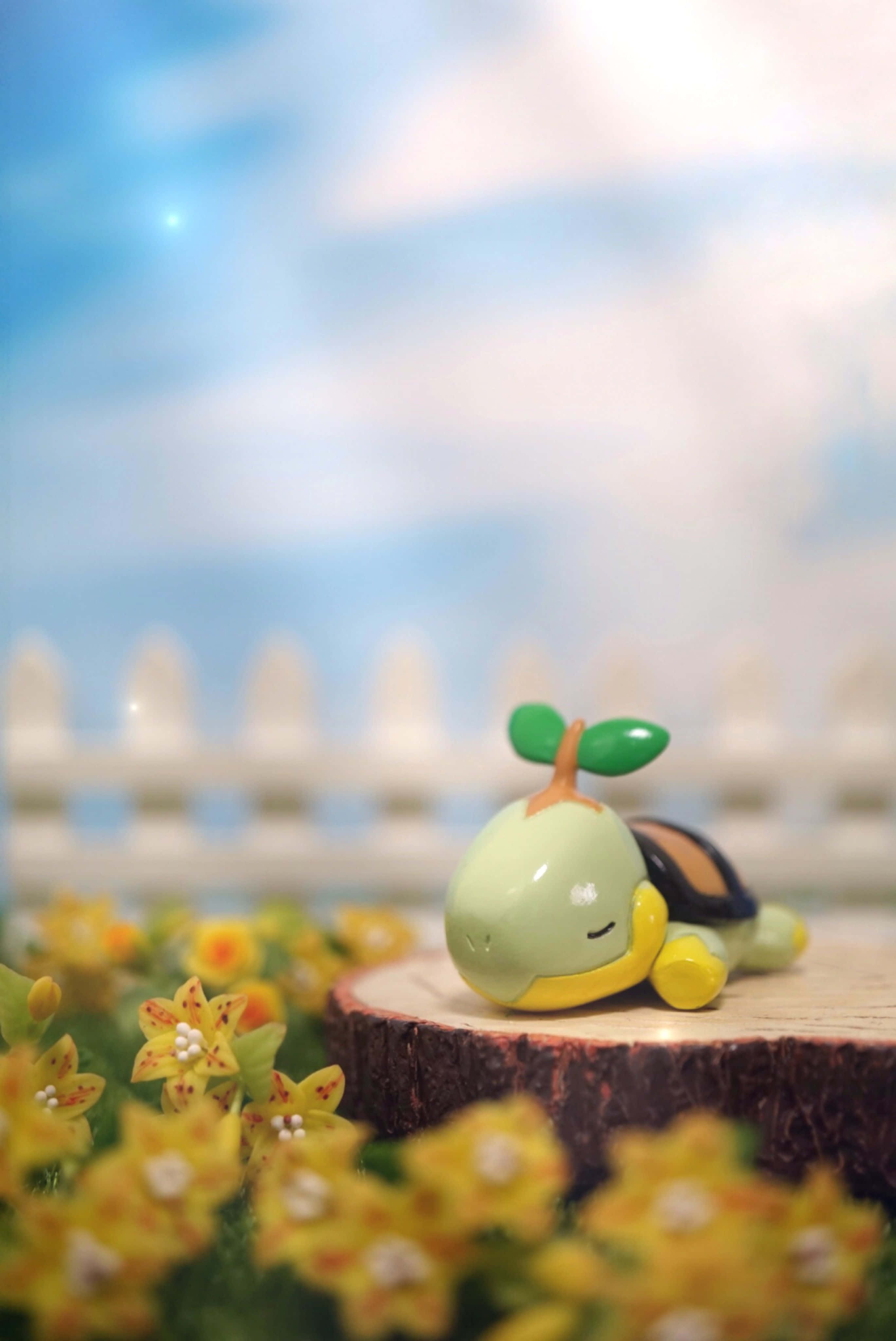 Pokemon Figures 桌布 壁紙 — 一個可愛的綠色烏龜雕像放在一個木樁上，周圍環繞著黃色花朵，在柔和的藍天下。