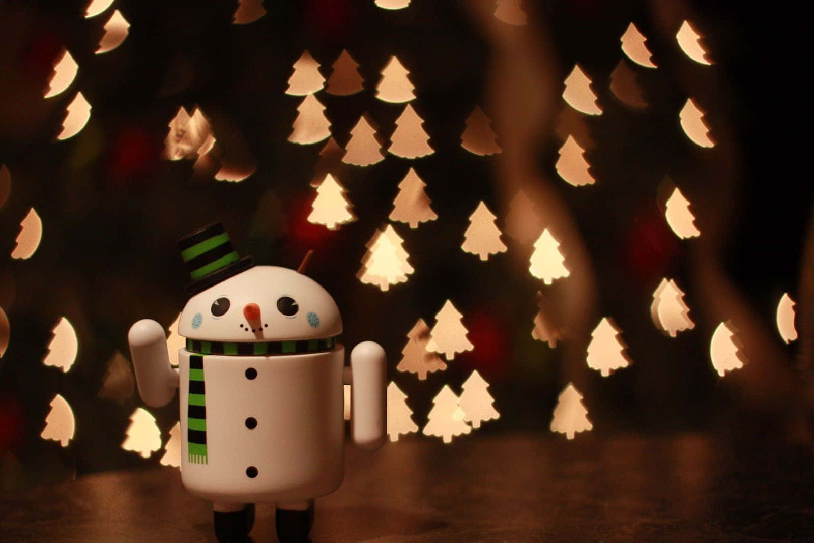 Tumblr Christmas 桌布 壁紙 — 一個可愛的雪人公仔，戴著綠色條紋圍巾和高帽，站在發光的散景聖誕樹燈光背景前。