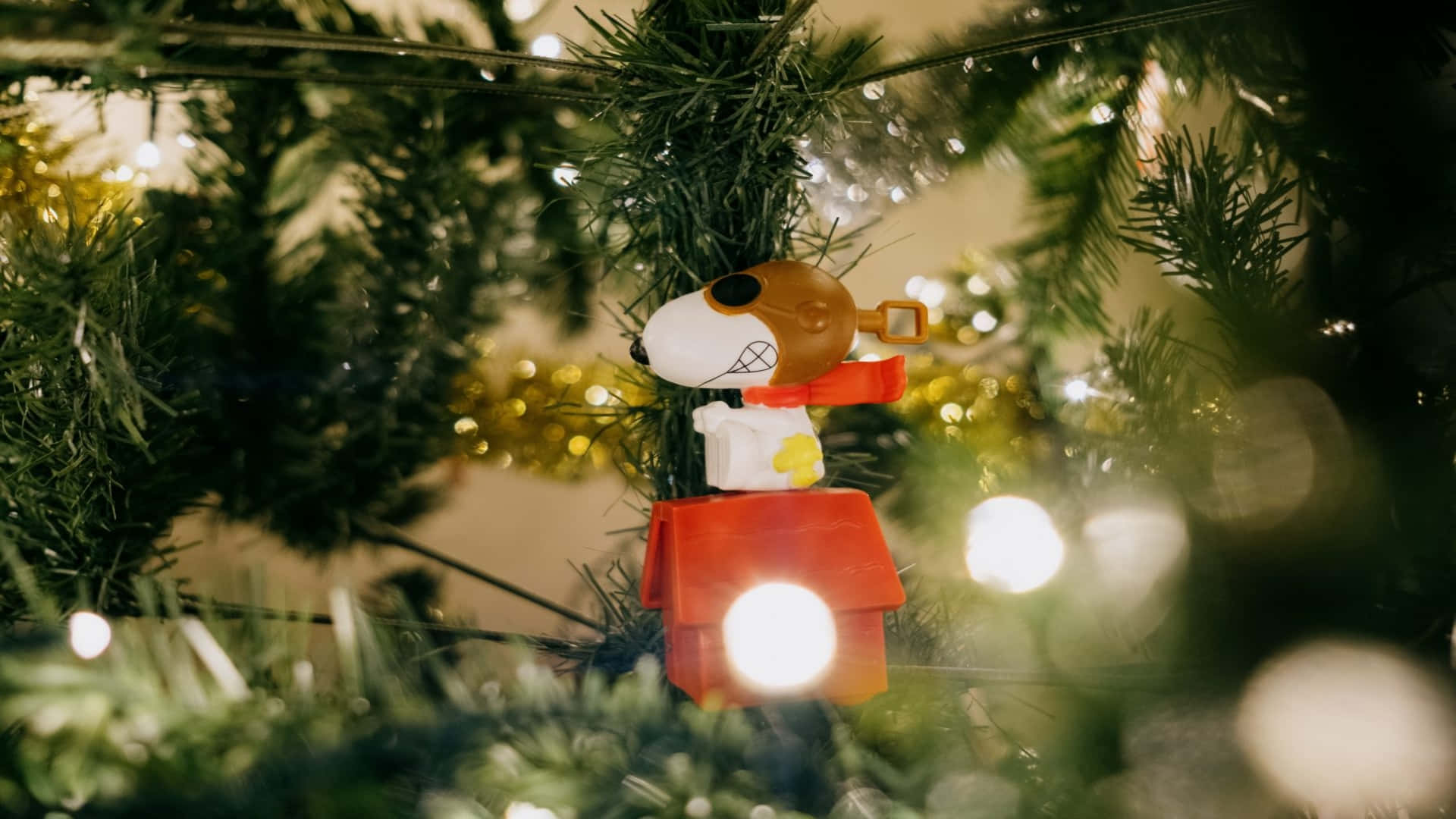 節日 桌布 壁紙 — 一個史努比在他紅色狗屋裡的人偶掛在一棵有閃爍燈光和綠色松針的聖誕樹上。