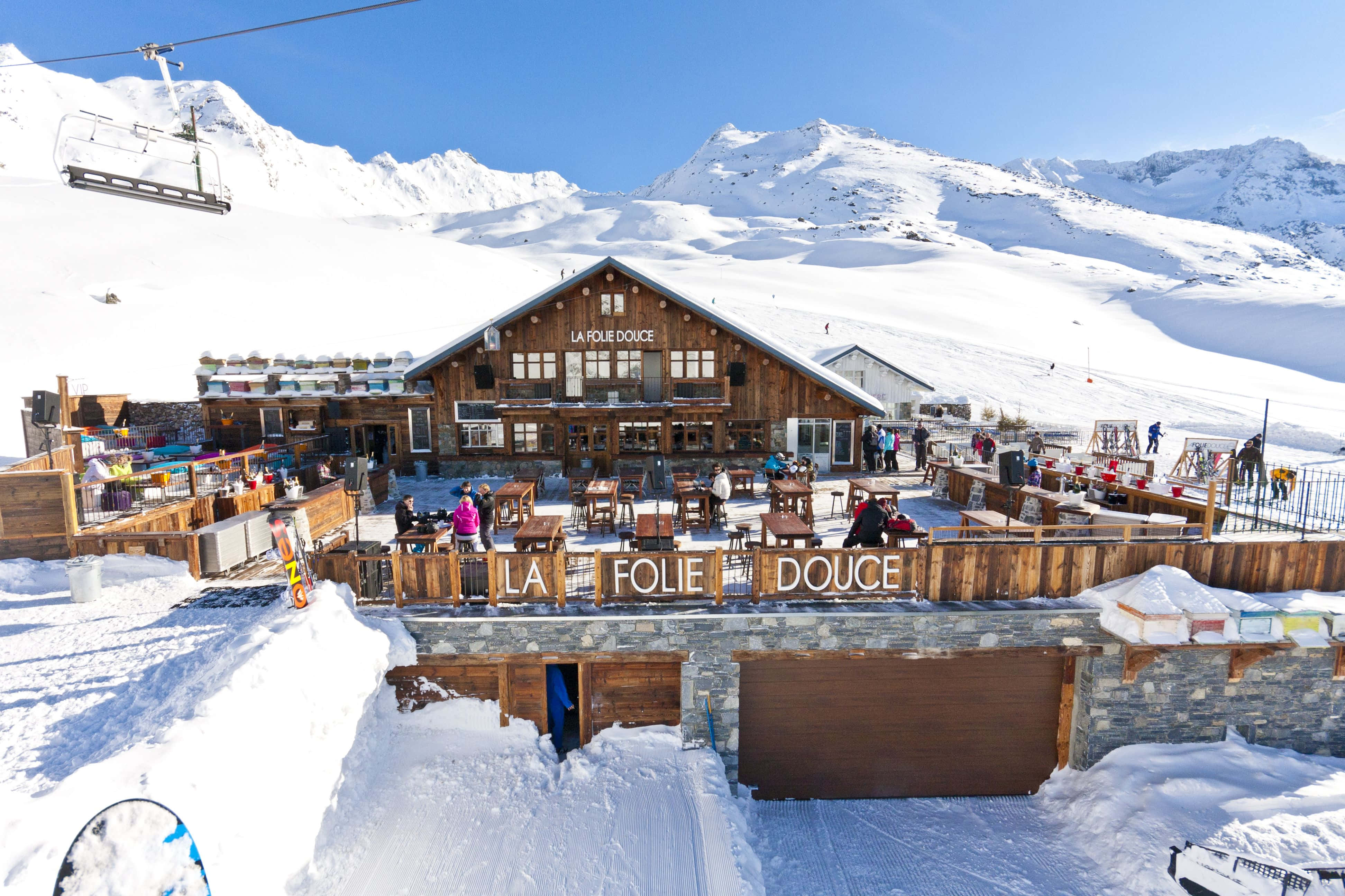 Ski Resort 桌布 壁紙 — 一個名為 La Folie Douce 的陽光滑雪小屋，坐落在白雪覆蓋的山上，背景有滑雪者和纜車。
