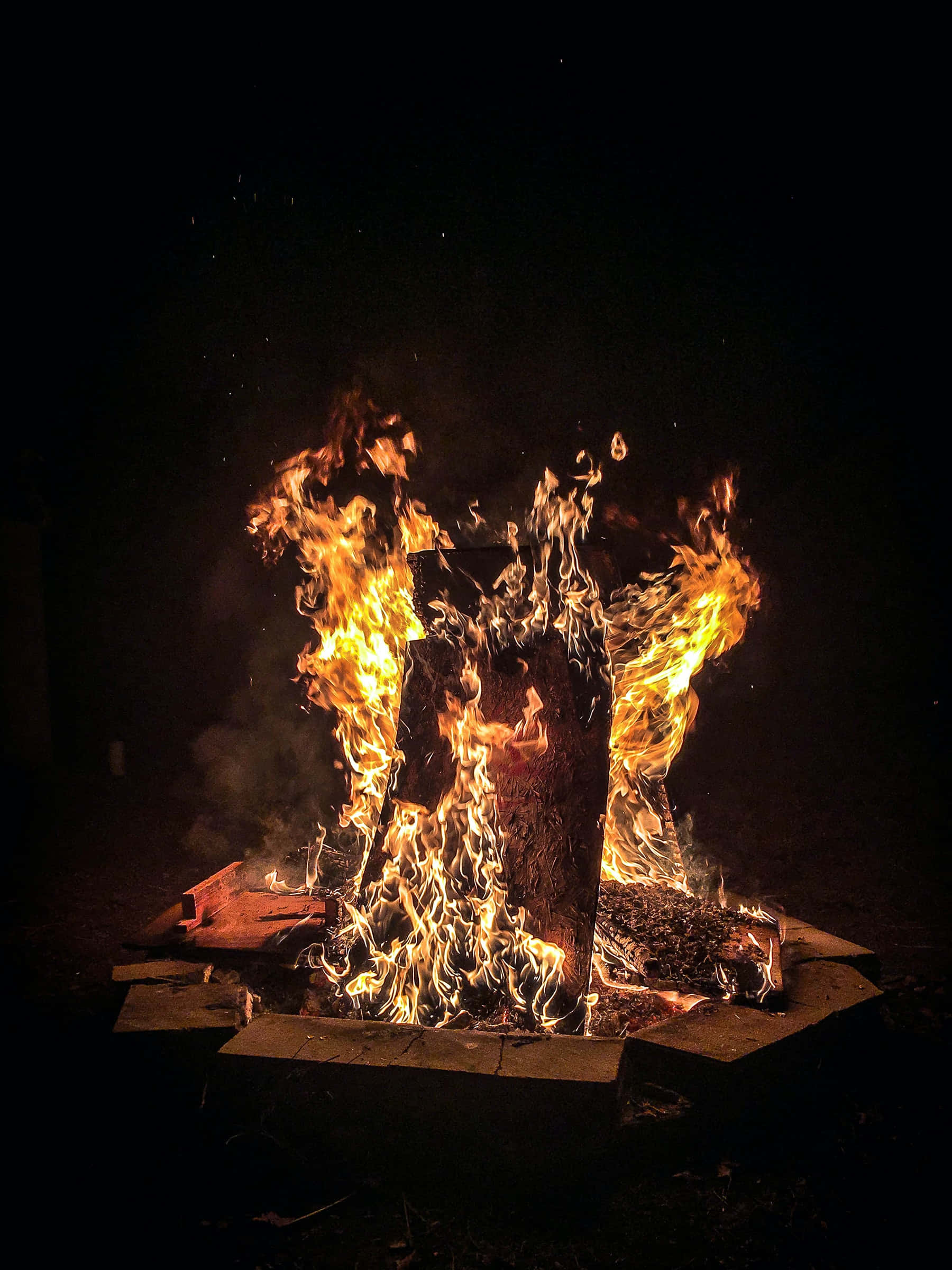Bonfire 桌布 壁紙 — 一個咆哮的營火在夜間明亮地燃燒，在黑暗的天空下，將金色的光芒投射在石頭火坑中的木頭上。