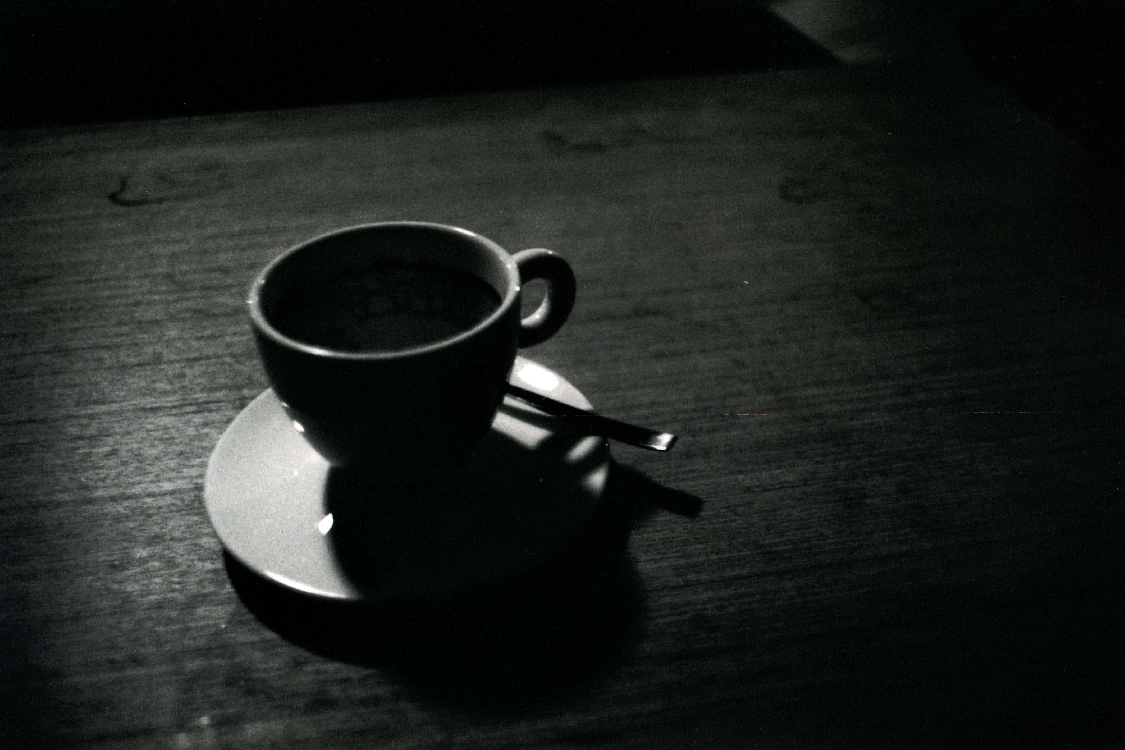 Black Coffee 桌布 壁紙 — 一個咖啡杯放在碟子上，擱在木桌上，在單色調的戲劇性聚光燈下。