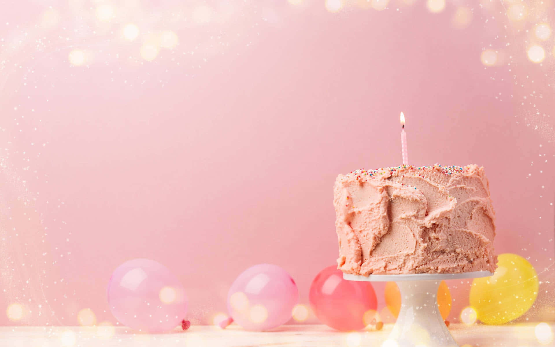 Birthday Cake 桌布 壁紙 — 一個單一的粉紅色糖霜生日蛋糕，上面有一根蠟燭，放在一個支架上，周圍環繞著彩色氣球，背景是柔和的粉紅色散景。