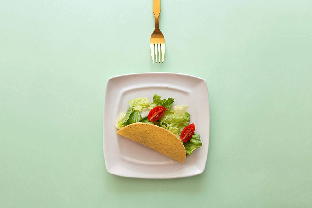 Taco 桌布 壁紙 — 一個單獨的墨西哥捲餅，配有生菜和番茄，放在白色盤子上，背景是薄荷綠色，上方有金色叉子。