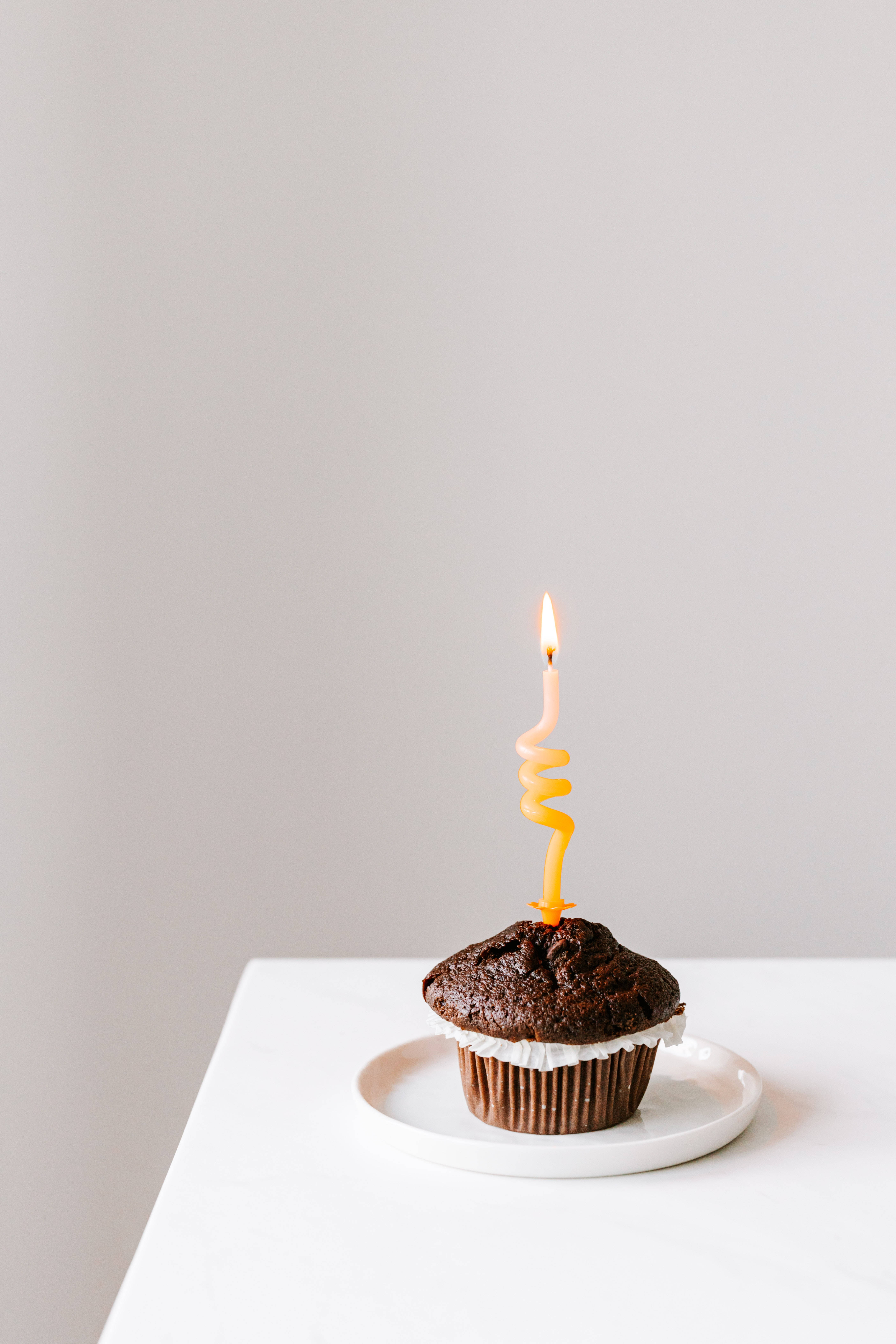 Birthday 桌布 壁紙 — 一個單獨的巧克力杯子蛋糕，上面有白色糖霜和一支點燃的黃色螺旋蠟燭，放在白色盤子上，背景是純淺灰色。