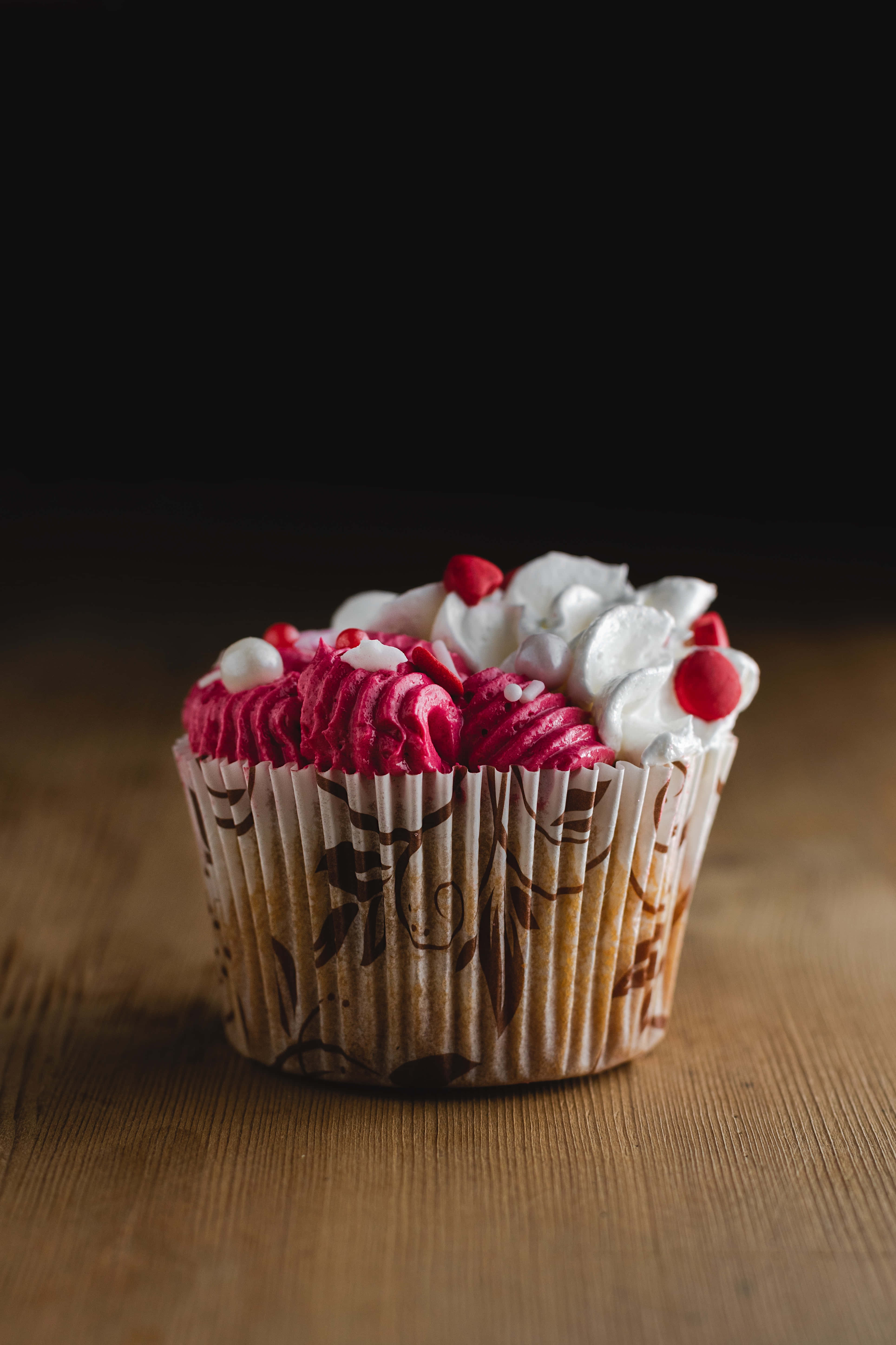 Cupcake 桌布 壁紙 — 一個單獨的杯子蛋糕，上面有粉紅色和白色的糖霜和紅色櫻桃配料，放在木質表面上，背景為深色。