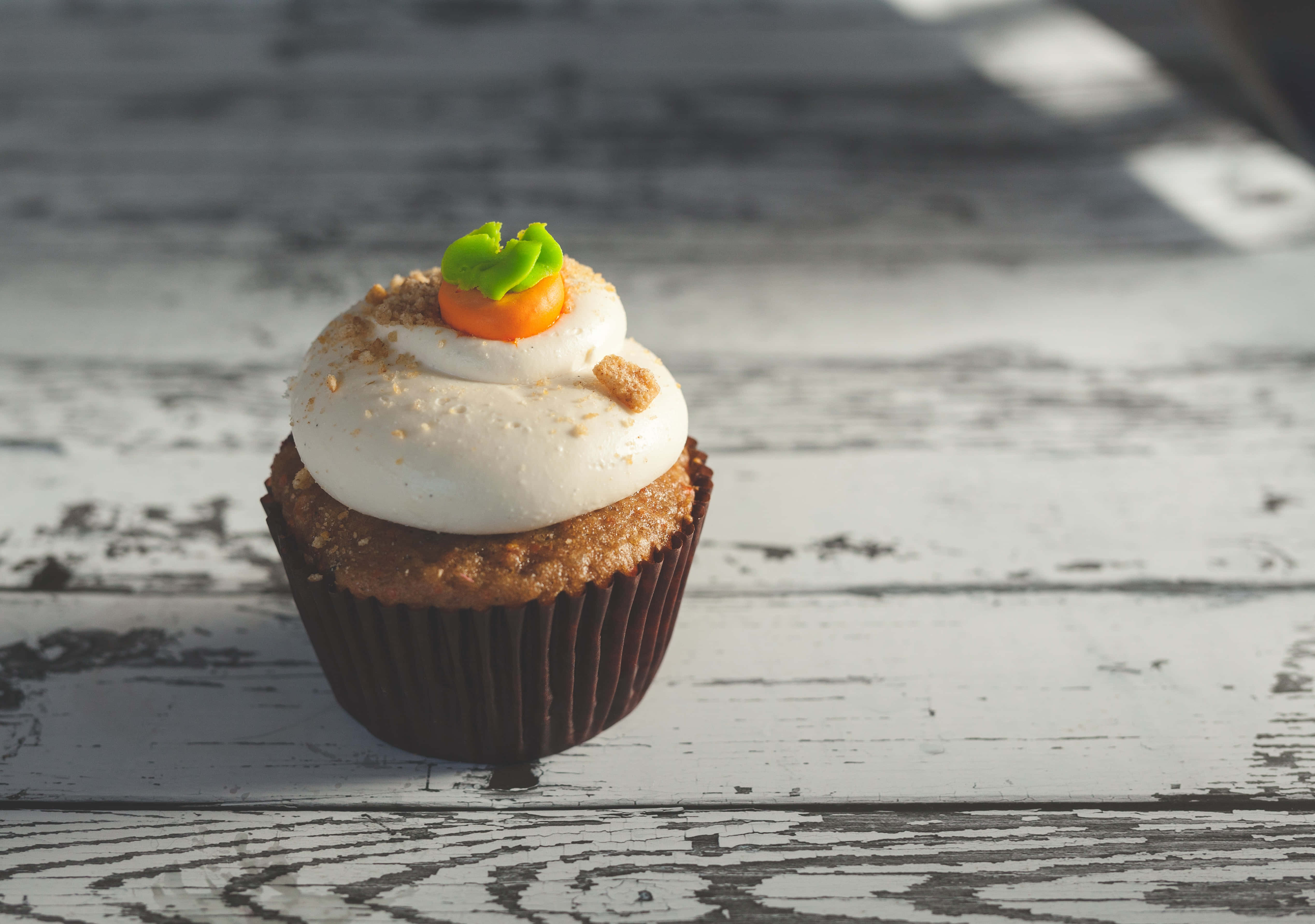 Cupcake 桌布 壁紙 — 一個單獨的胡蘿蔔蛋糕杯子蛋糕，上面有白色糖霜和胡蘿蔔裝飾，放在一個風化的白色木質表面上。