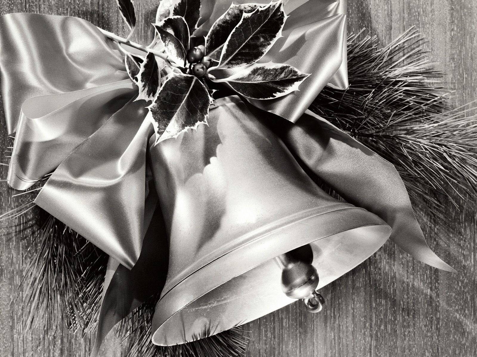 Christmas Bell 桌布 壁紙 — 一個單色特寫，展示一個閃亮的銀色鈴鐺，飾有冬青漿果和松針，用緞帶蝴蝶結綁在木質表面上。