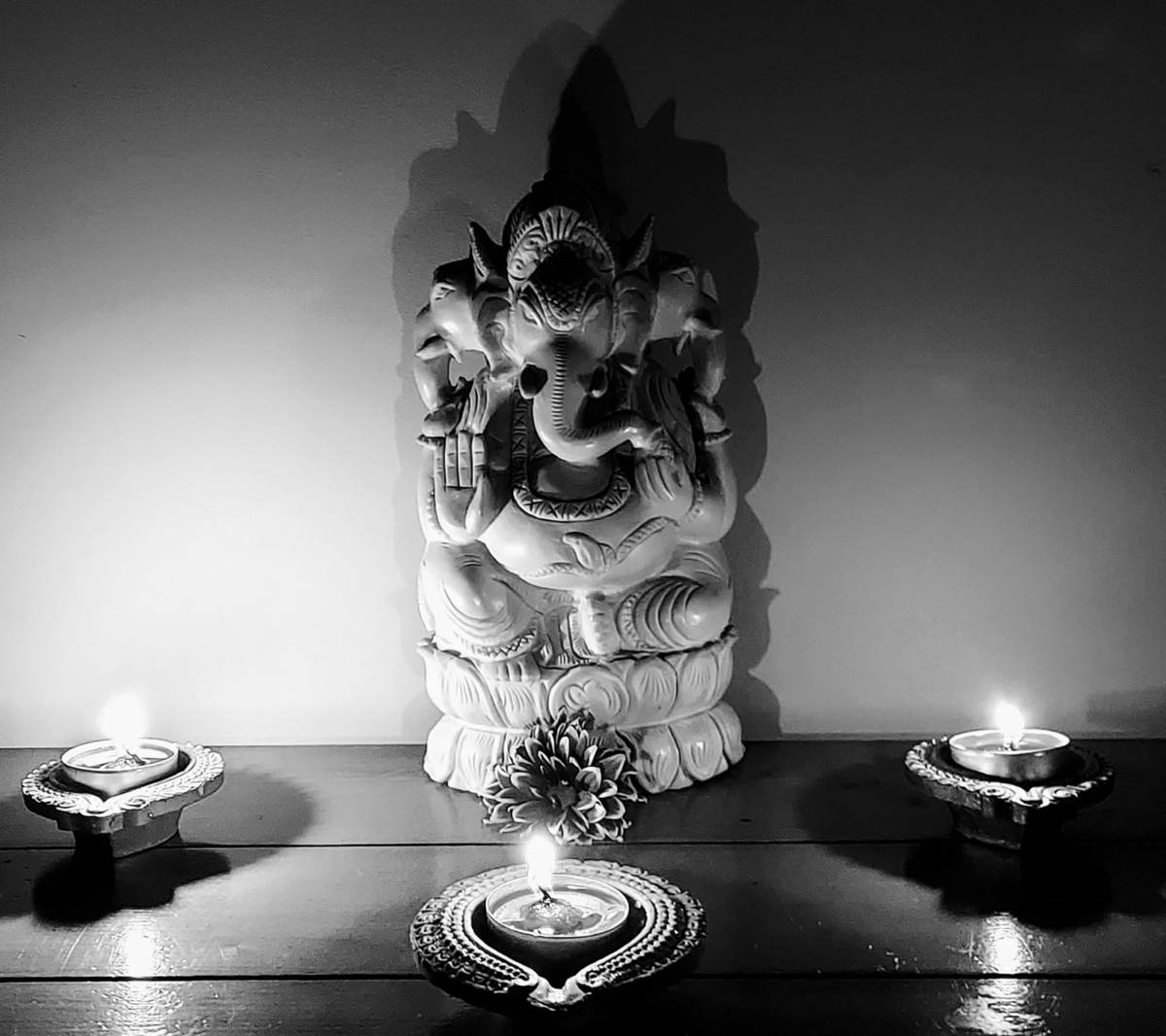 Ganesh 桌布 壁紙 — 一個單色象頭神雕像安詳地坐在木質表面上，兩側有三盞點燃的油燈，投下柔和陰影。