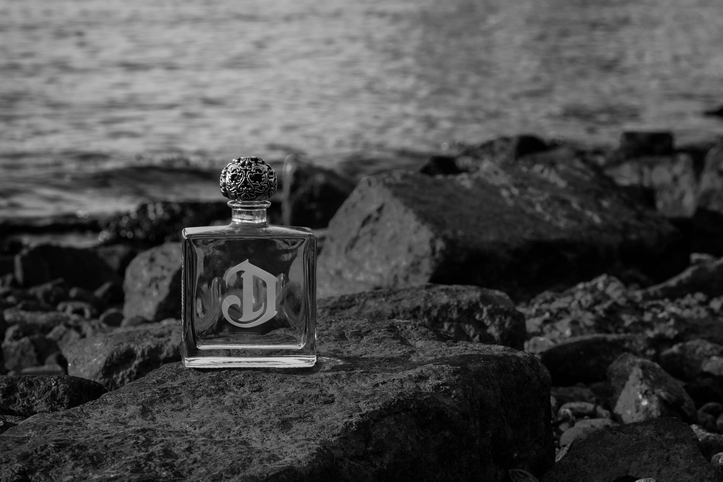 alcohol 桌布 壁紙 — 一個單色香水瓶，帶有華麗的瓶蓋和「D」字母組合，放置在崎嶇的岩石上，旁邊是模糊的水岸線。