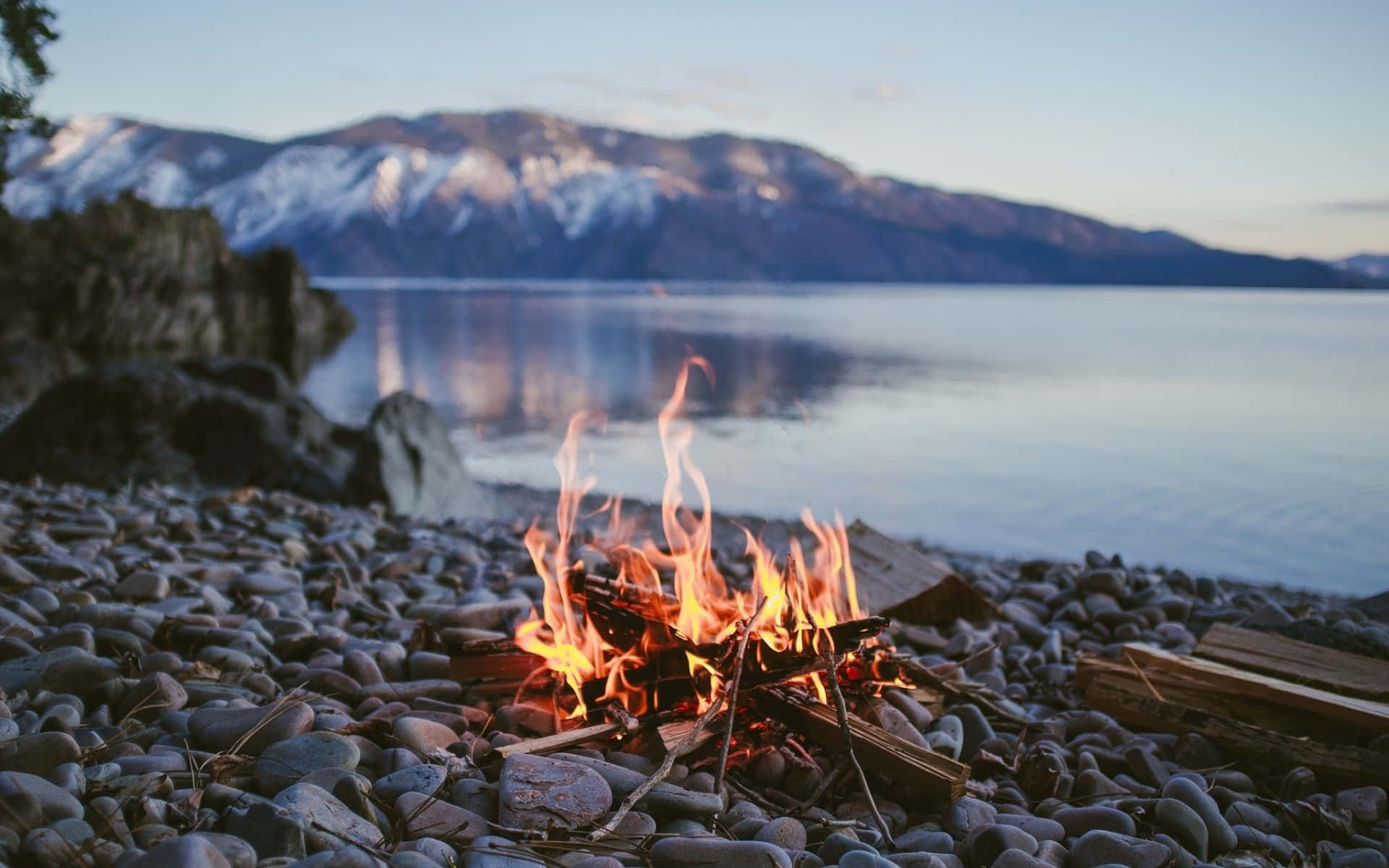 Beach Bonfire 桌布 壁紙 — 一個噼啪作響的營火在佈滿鵝卵石的湖岸上明亮地燃燒，白雪覆蓋的山脈在黃昏時分倒映在平靜的水面上。