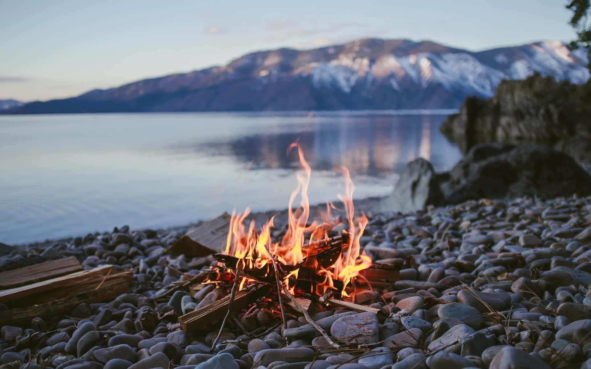 Camping 桌布 壁紙 — 一個噼啪作響的營火在岩石湖岸上明亮地燃燒，積雪覆蓋的山脈倒映在黃昏平靜的水面上。