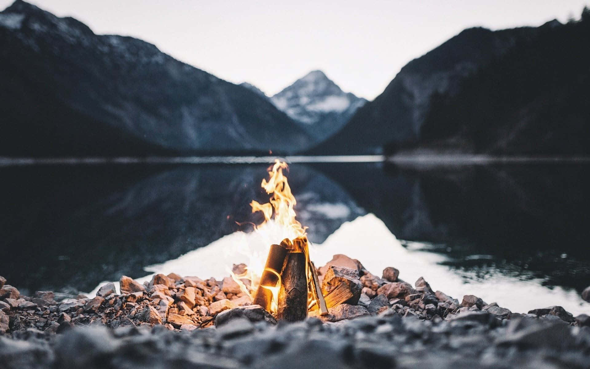 Beach Bonfire 桌布 壁紙 — 一個噼啪作響的營火在岩石湖岸上溫暖地發光，白雪覆蓋的山脈倒映在黃昏平靜的水面上。