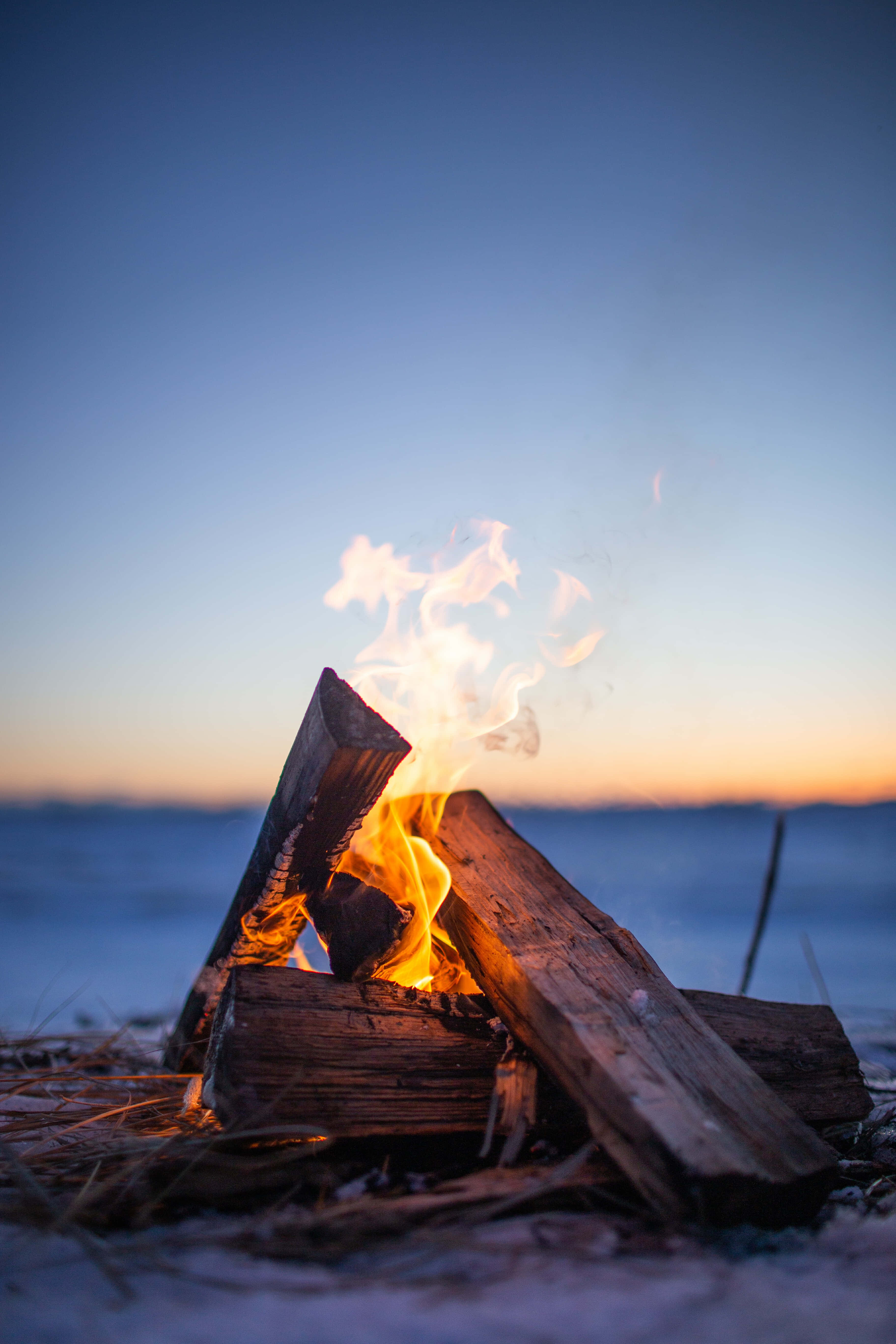 Camping 桌布 壁紙 — 一個噼啪作響的營火在黃昏時分在白雪覆蓋的海灘上明亮地燃燒，將溫暖的光芒投射在清冷的藍天上。